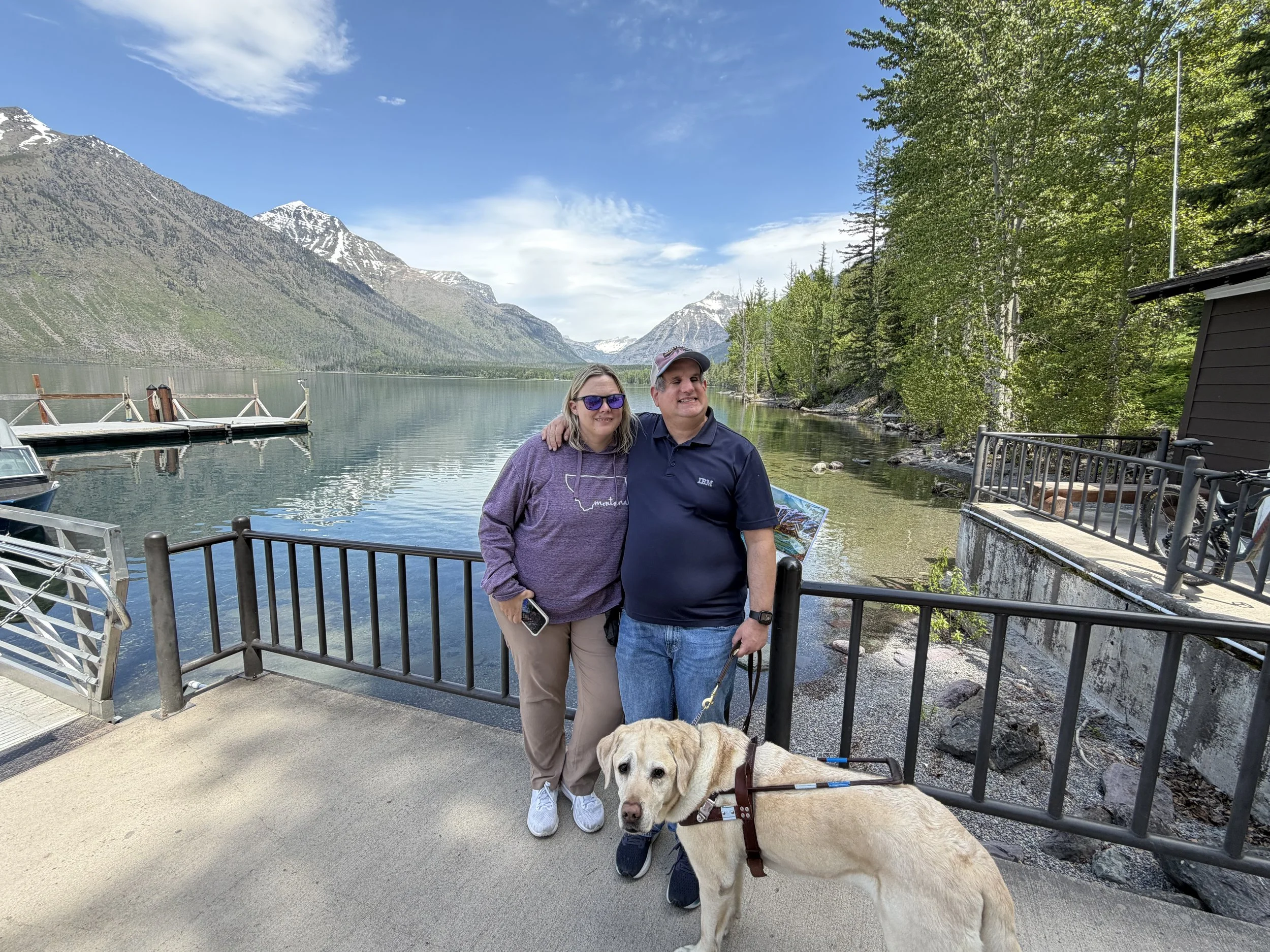 Two people standing on a dock by a lake with mountains in the background, accompanied by a yellow Labrador retriever.