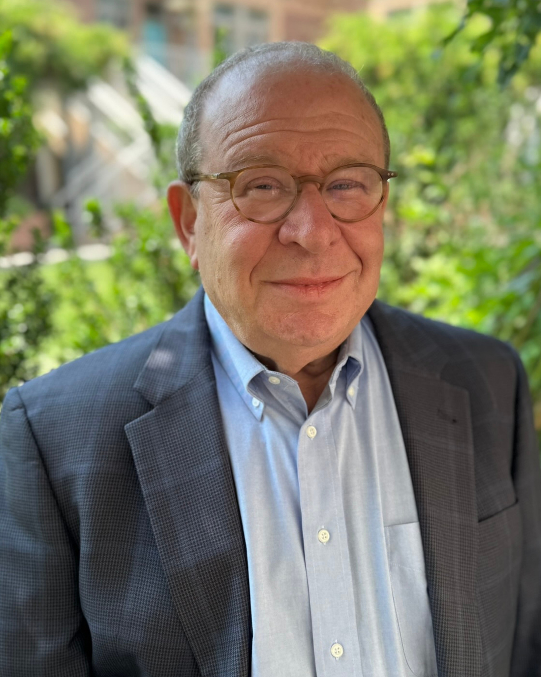 A smiling middle-aged man wearing glasses, a blue dress shirt, and a dark blazer, standing outdoors with green foliage in the background.