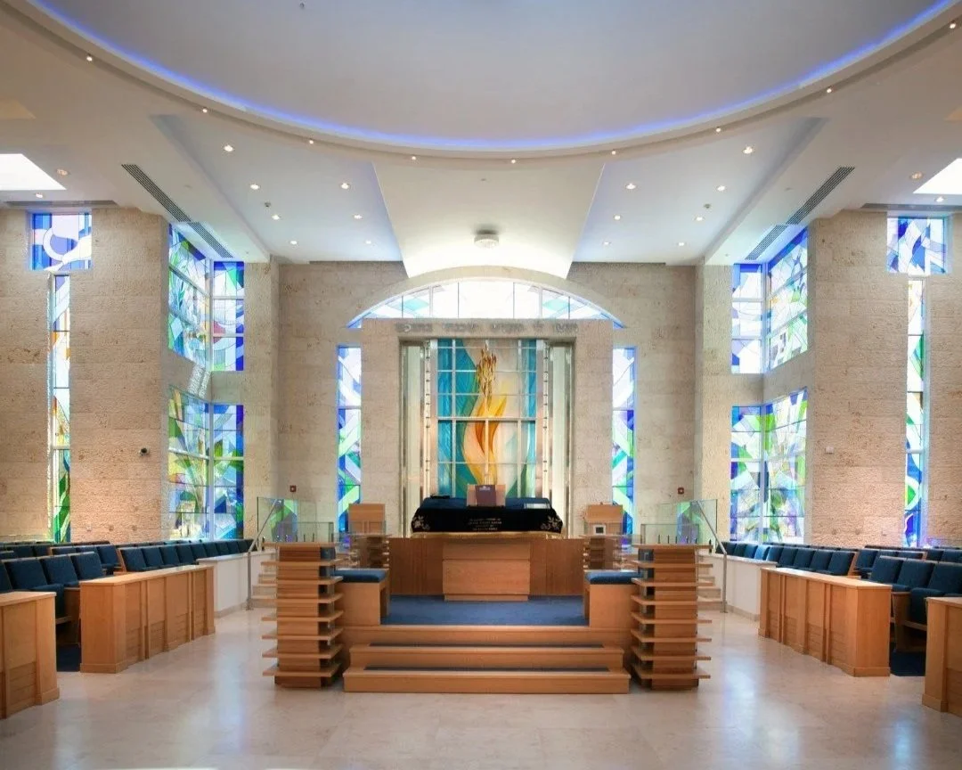 Interior of a church with wooden pews arranged on either side of a stage, stained glass windows on the walls, and a central altar with a colorful abstract artwork behind it, under a curved ceiling with recessed lighting.