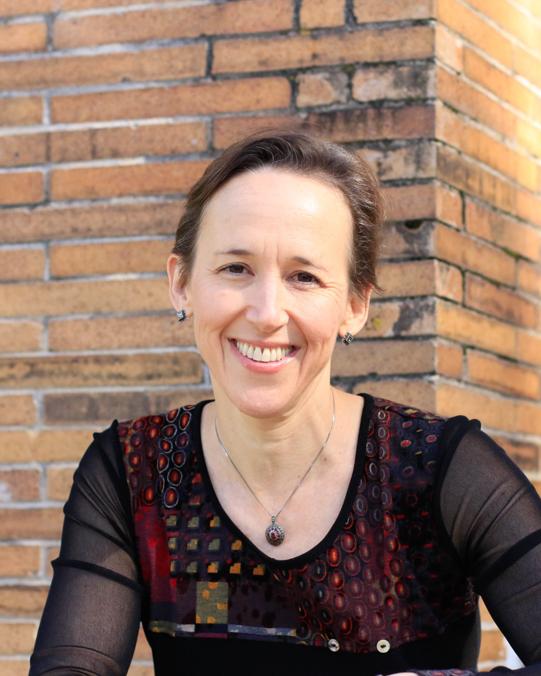 A smiling woman with short brown hair and earrings, wearing a black top with colorful patterns, sitting in front of a brick wall.