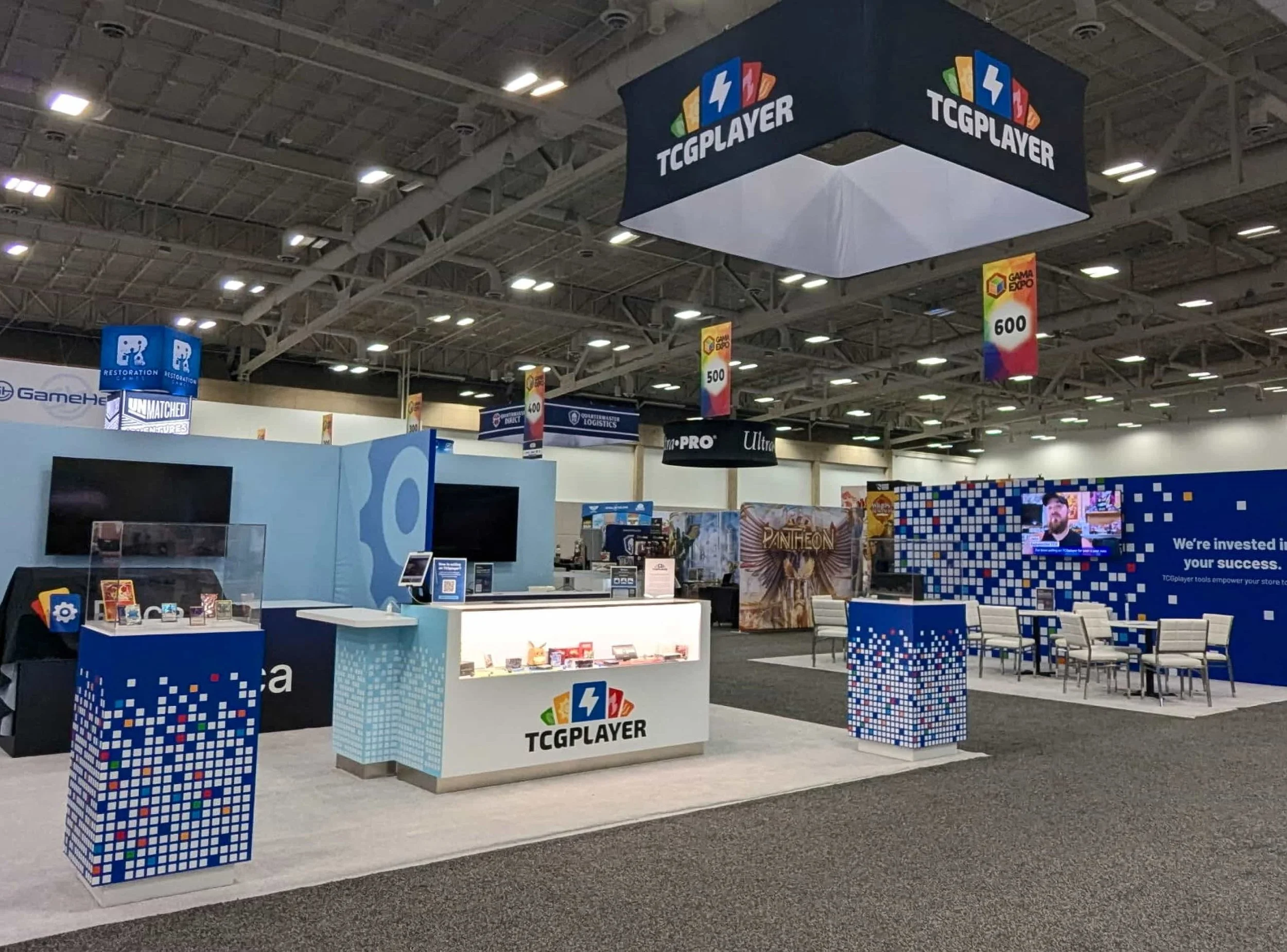 Live event photography of previously shown trade show booth, featuring white counter displaying merchandise, colorful pixelated panels on the sides, and hanging signs with numbers and the GAMA Expo logo in a large exhibition hall.