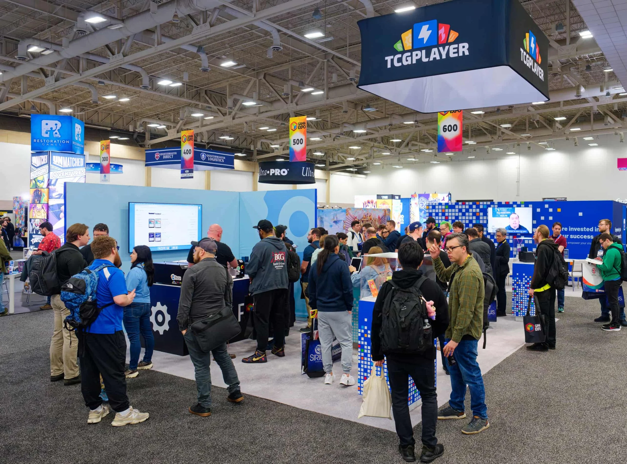 An indoor convention or expo with many attendees walking and standing around booths and displays. Oversized hanging signs indicate booth numbers 300, 400, 500, and 600. The image shows a crowd of people, some wearing backpacks, and various company or event banners, such as GAMA Expo and Restoration Games. The ceiling has exposed beams and lighting fixtures.