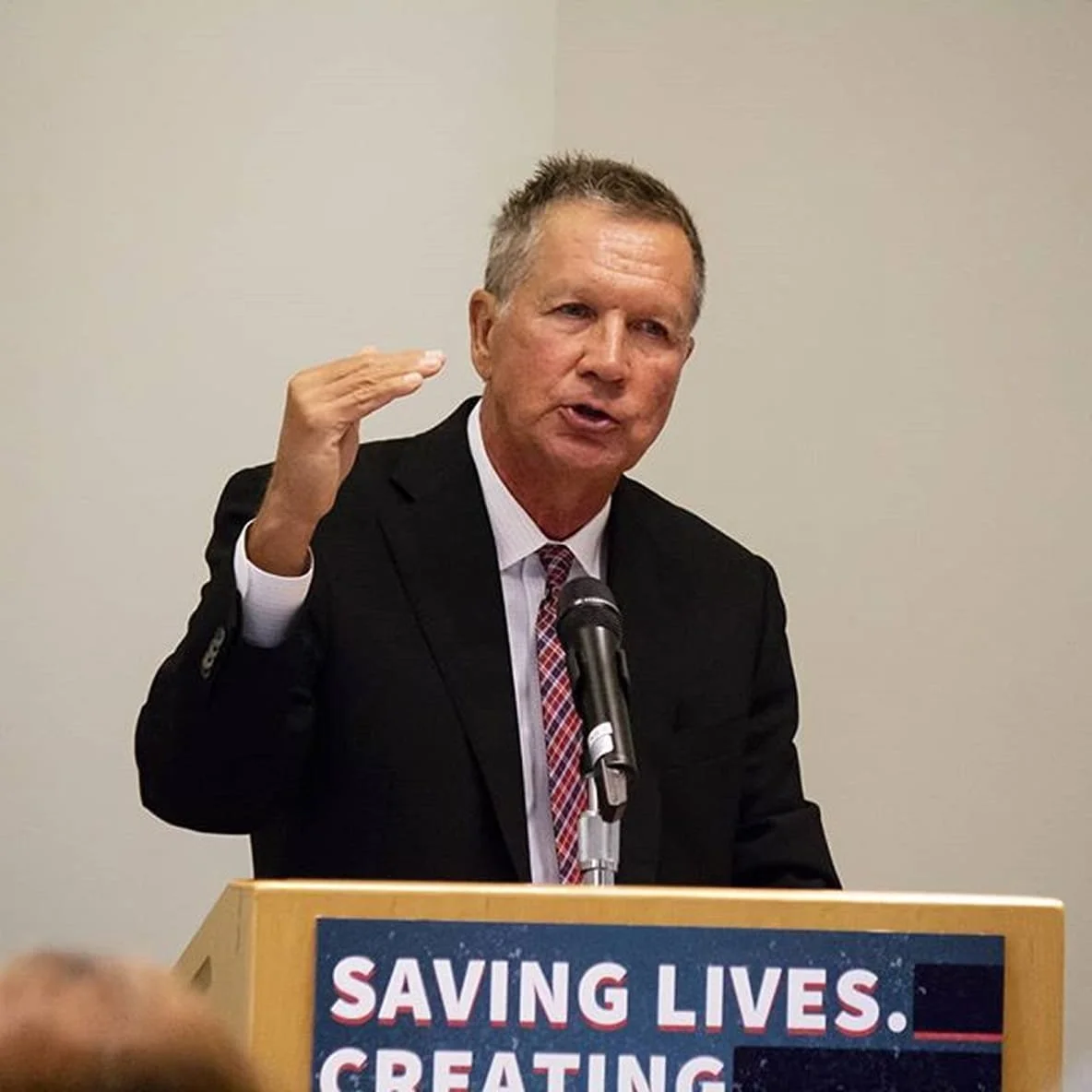 Former Ohio Governor John Kasich in a suit giving a speech behind a podium at an August 2018 press event.