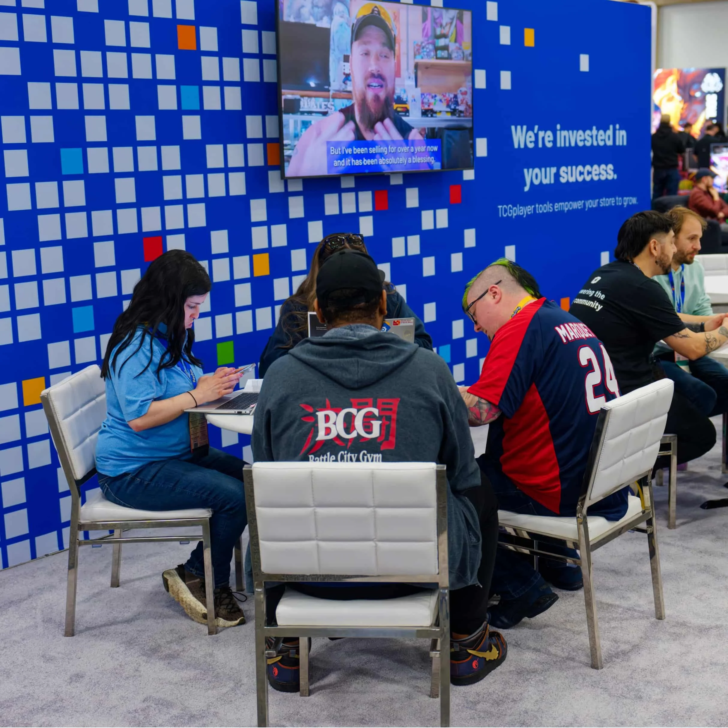 A group of people sitting at a table in TCGplayer's themed booth experience. The rear wall features a colorful pixel and a mounted television screen.