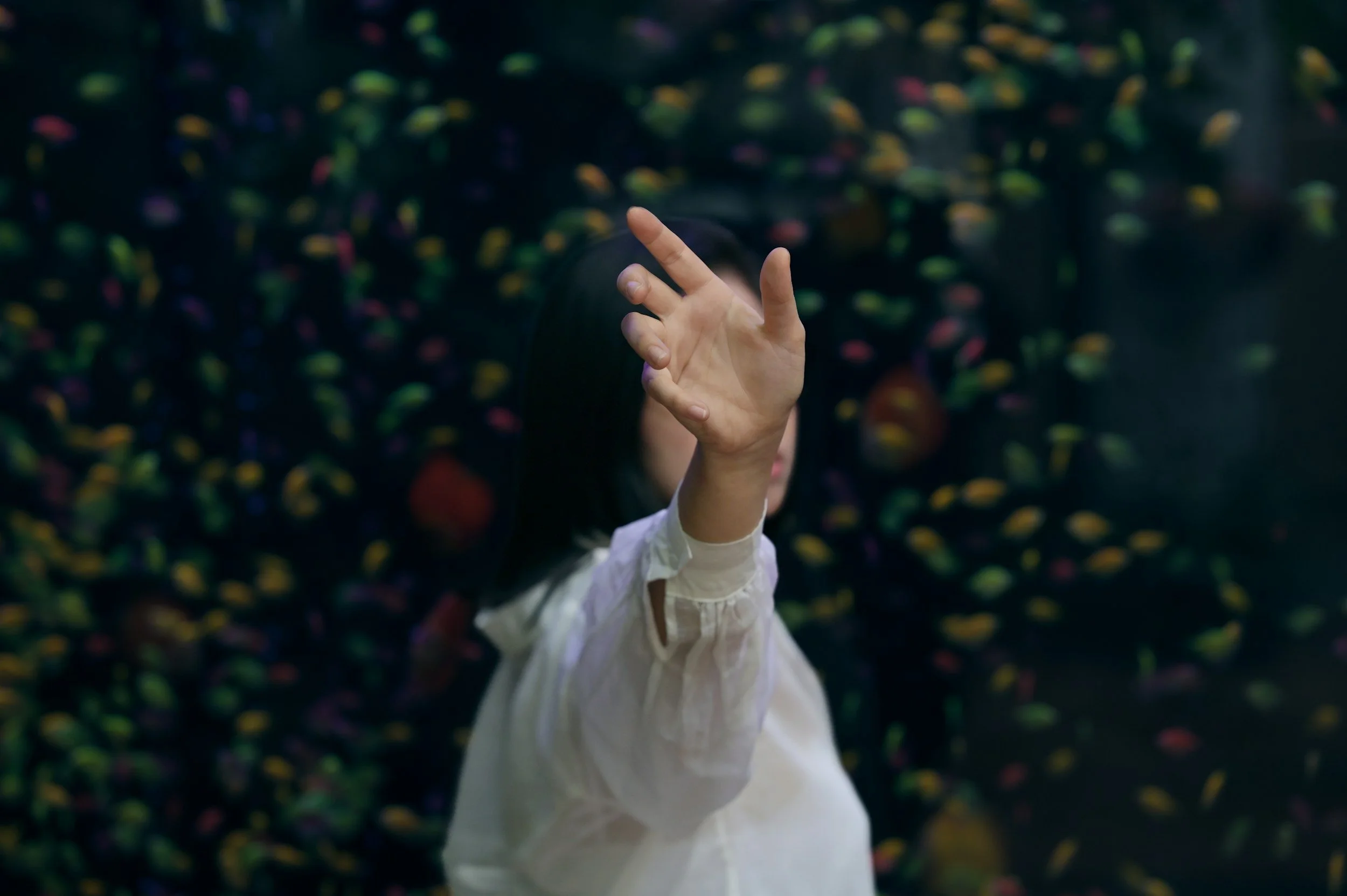 A child wearing a white shirt with a ruffled sleeve is reaching out with one finger. The background is filled with colorful bouncy balls or similar objects, creating a vibrant and playful atmosphere.