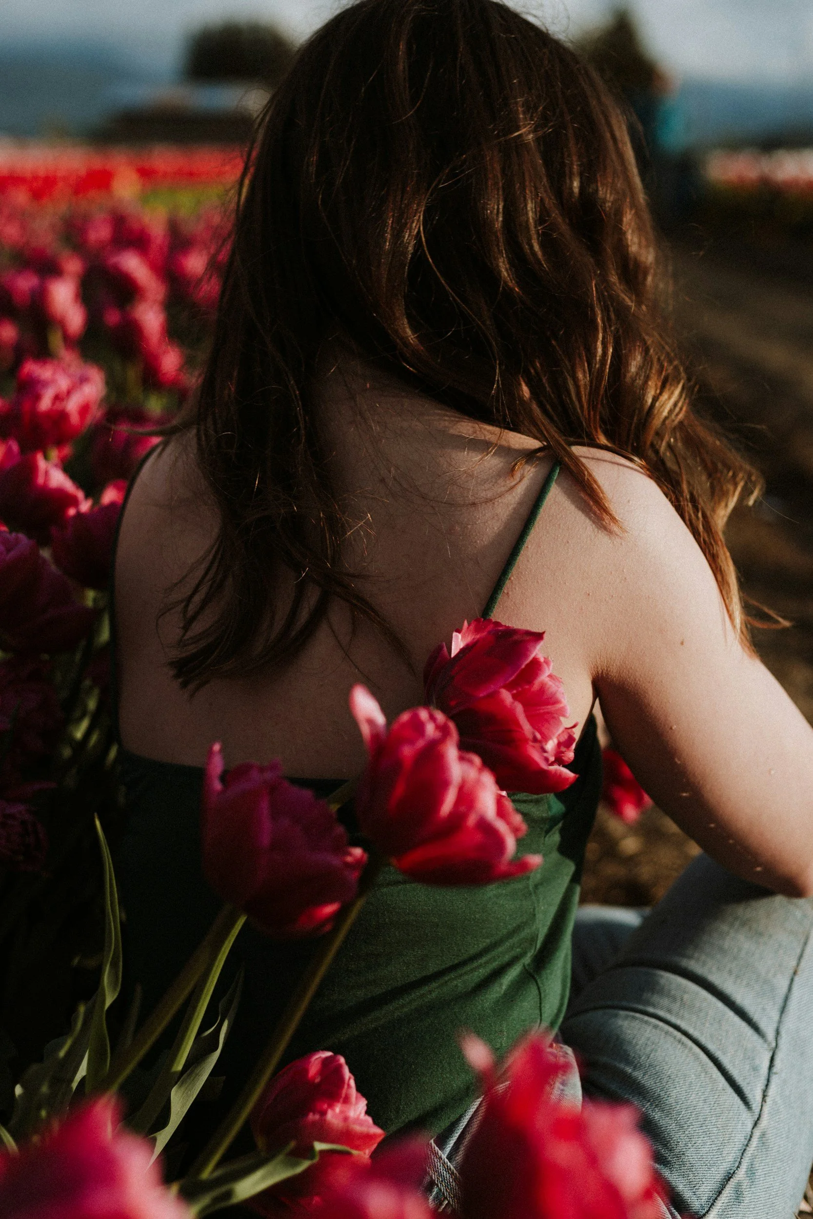 A woman with brown hair and a green top is sitting outdoors, surrounded by pink flowers. The scene is set during the evening with a dimly lit background.