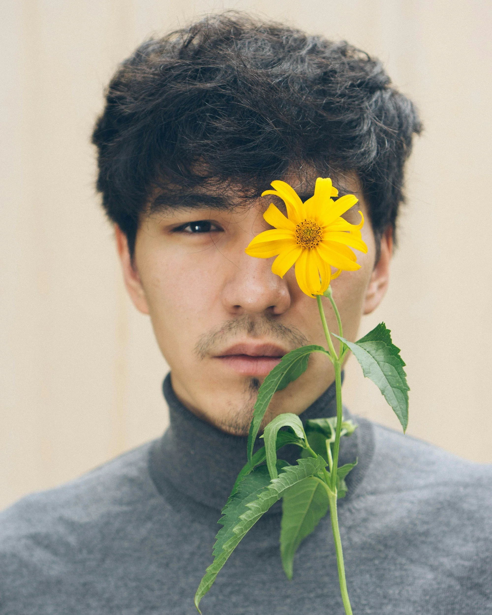 A young man with dark, wavy hair and a slight beard holds a yellow flower close to his face, with part of his face visible and a neutral background.