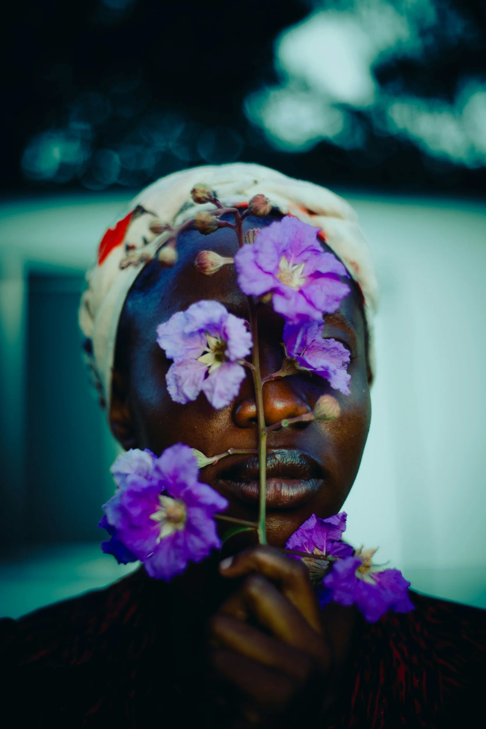 A portrait of a person with dark skin, closed eyes, adorned with purple flowers and a floral headpiece, against a blurred background.