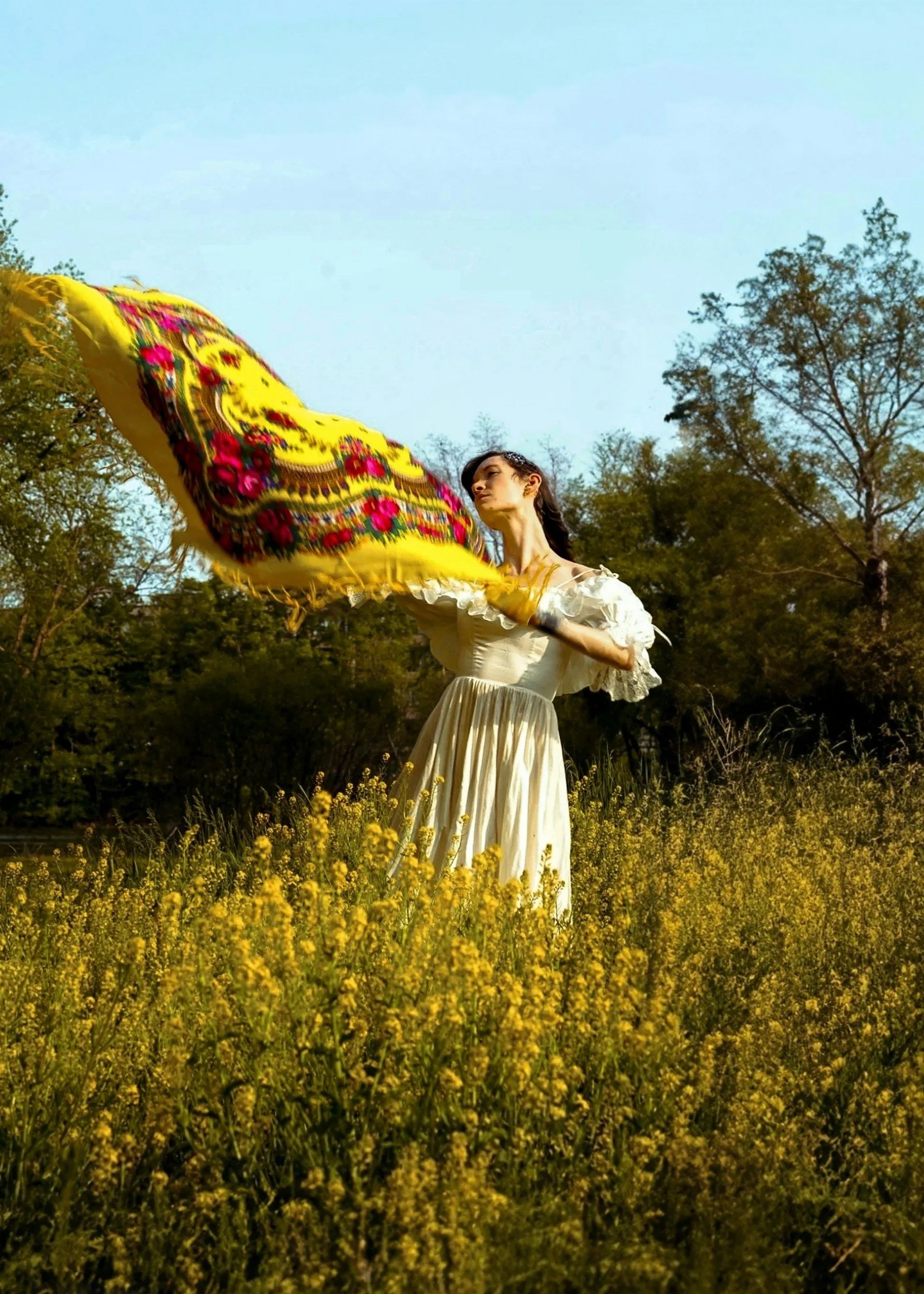 A woman in a white vintage dress standing in a yellow flower field, holding a large, vibrant yellow embroidered scarf, with trees and a clear blue sky in the background.