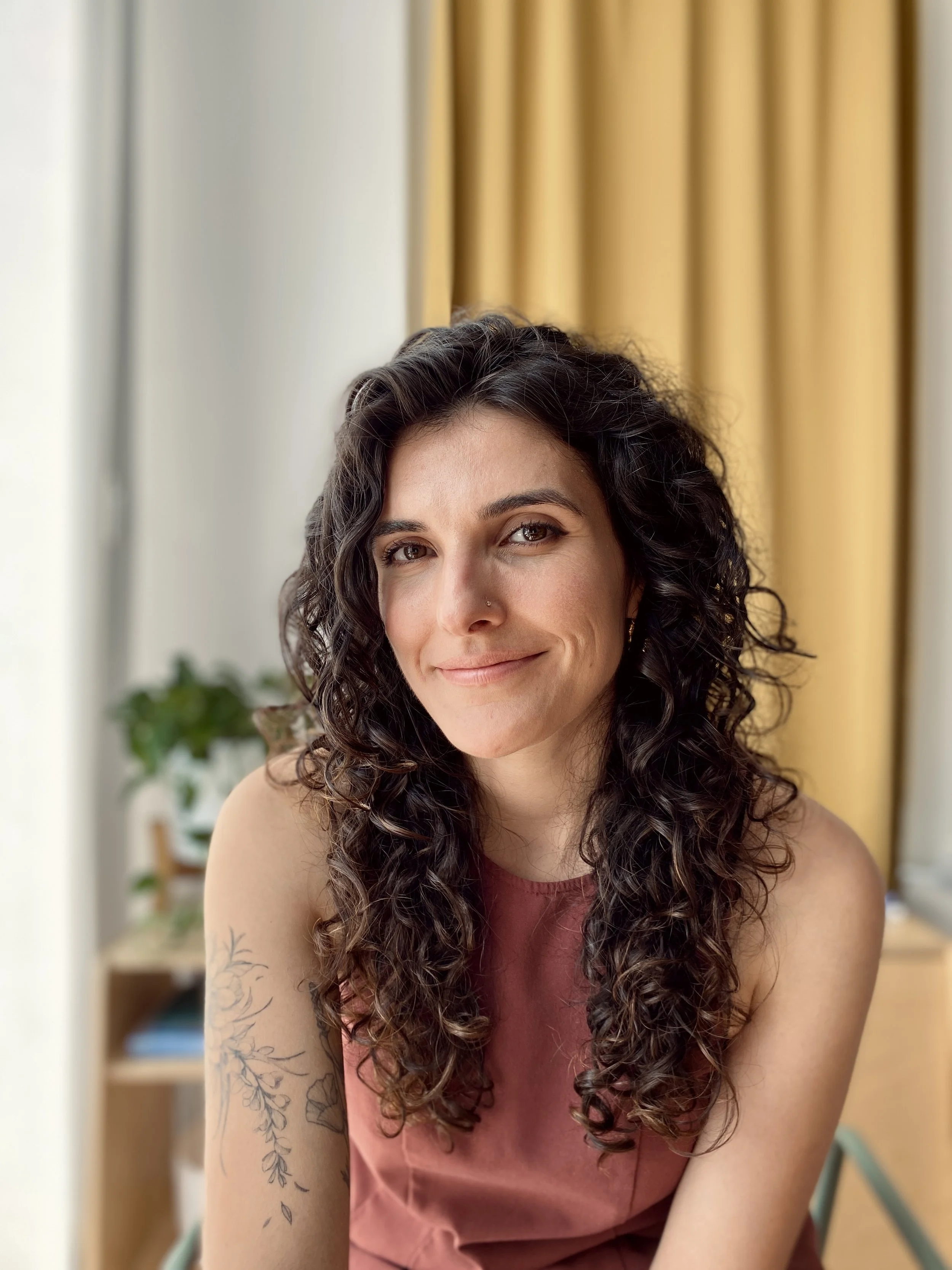 A woman with curly dark hair smiling in front of yellow curtain and blurred background.