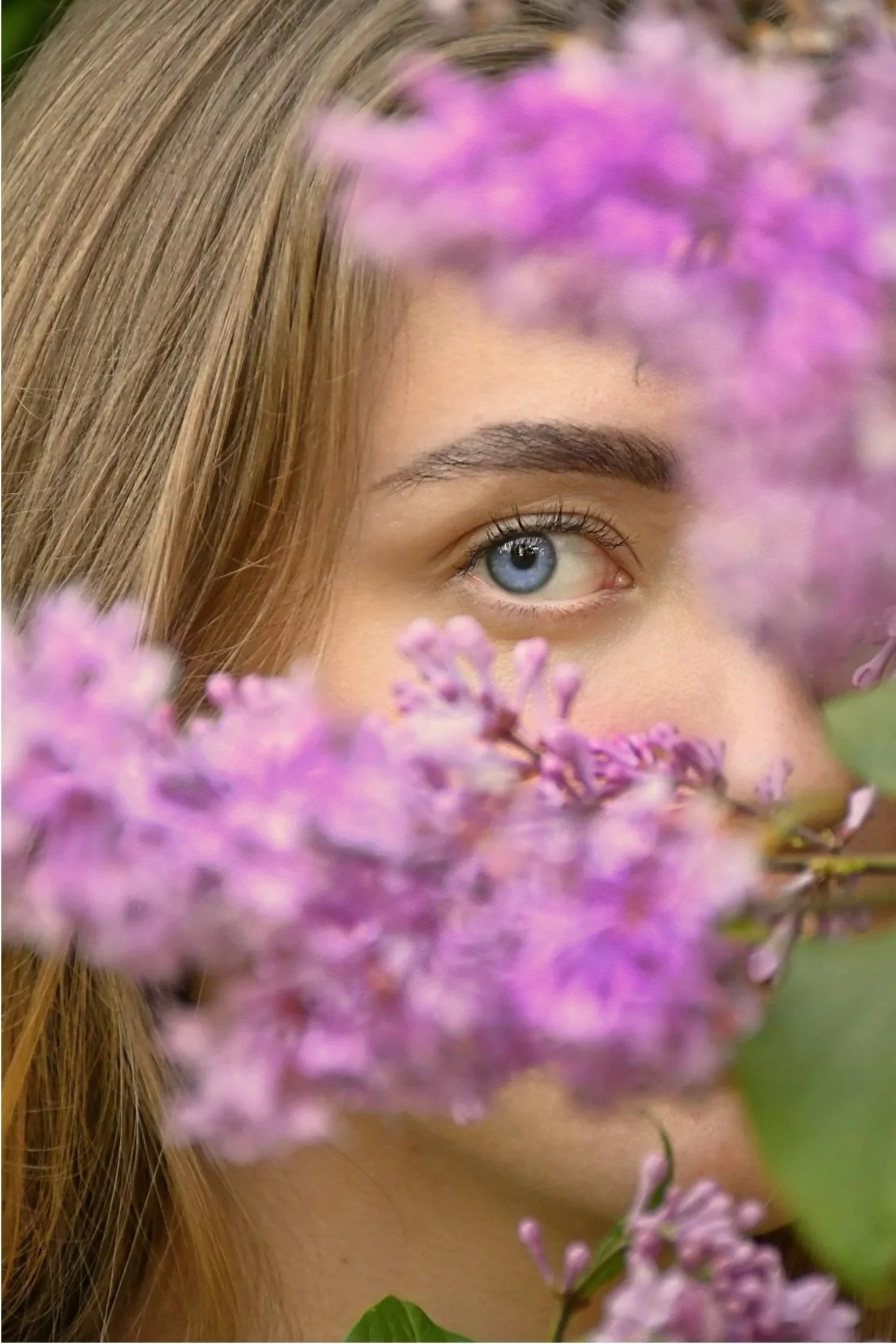 Close-up of a woman's face with only her eye and a part of her forehead visible, surrounded by purple flowers, with blonde hair partially covering her face.