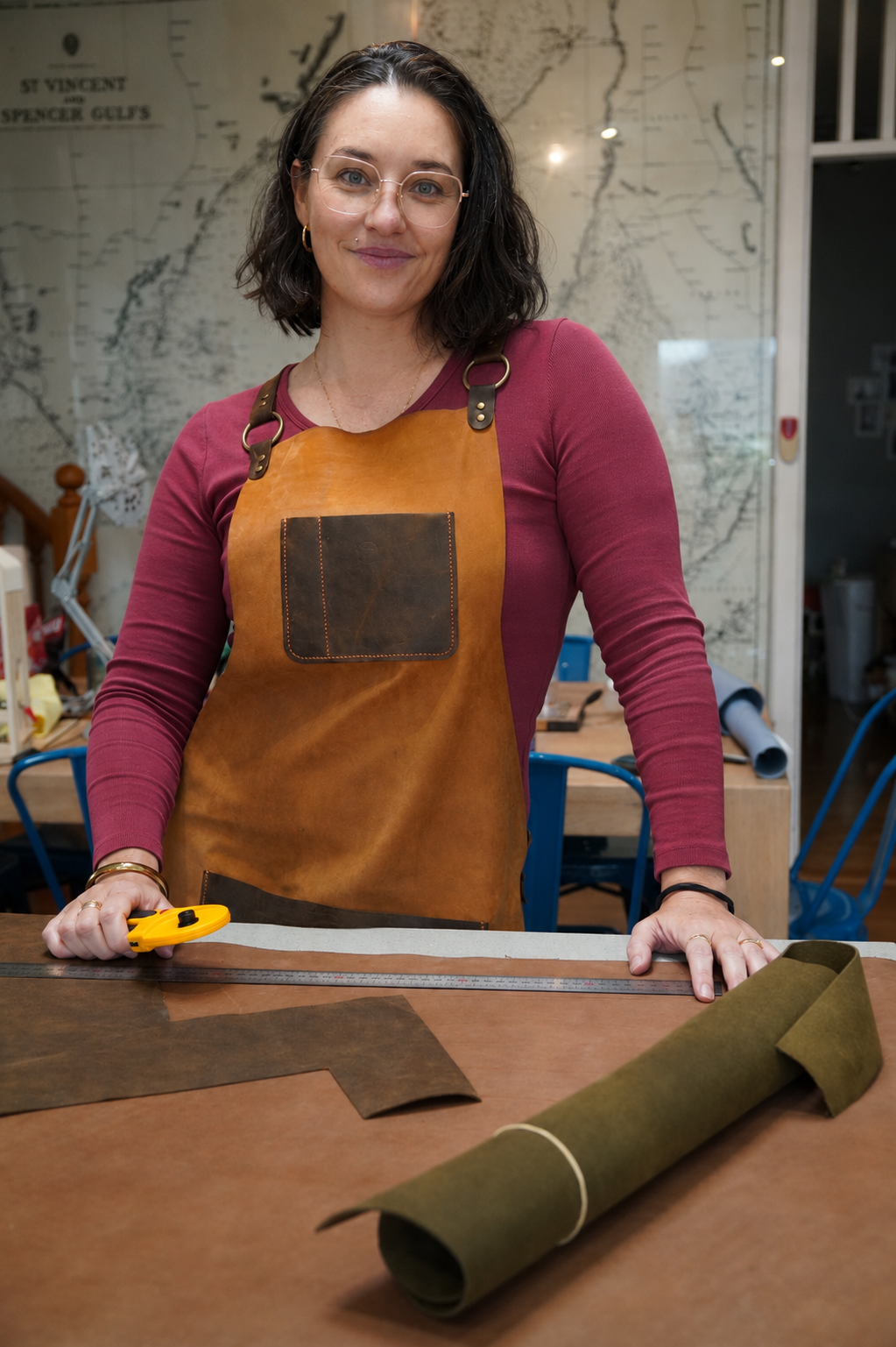 A woman wearing glasses, a maroon long-sleeved shirt, and a brown leather apron, stands at a workbench in a workshop. She is holding a yellow utility knife and a metal ruler, with a rolled piece of green fabric or felt and cut pieces on the table. A large map is hanging on the wall in the background.