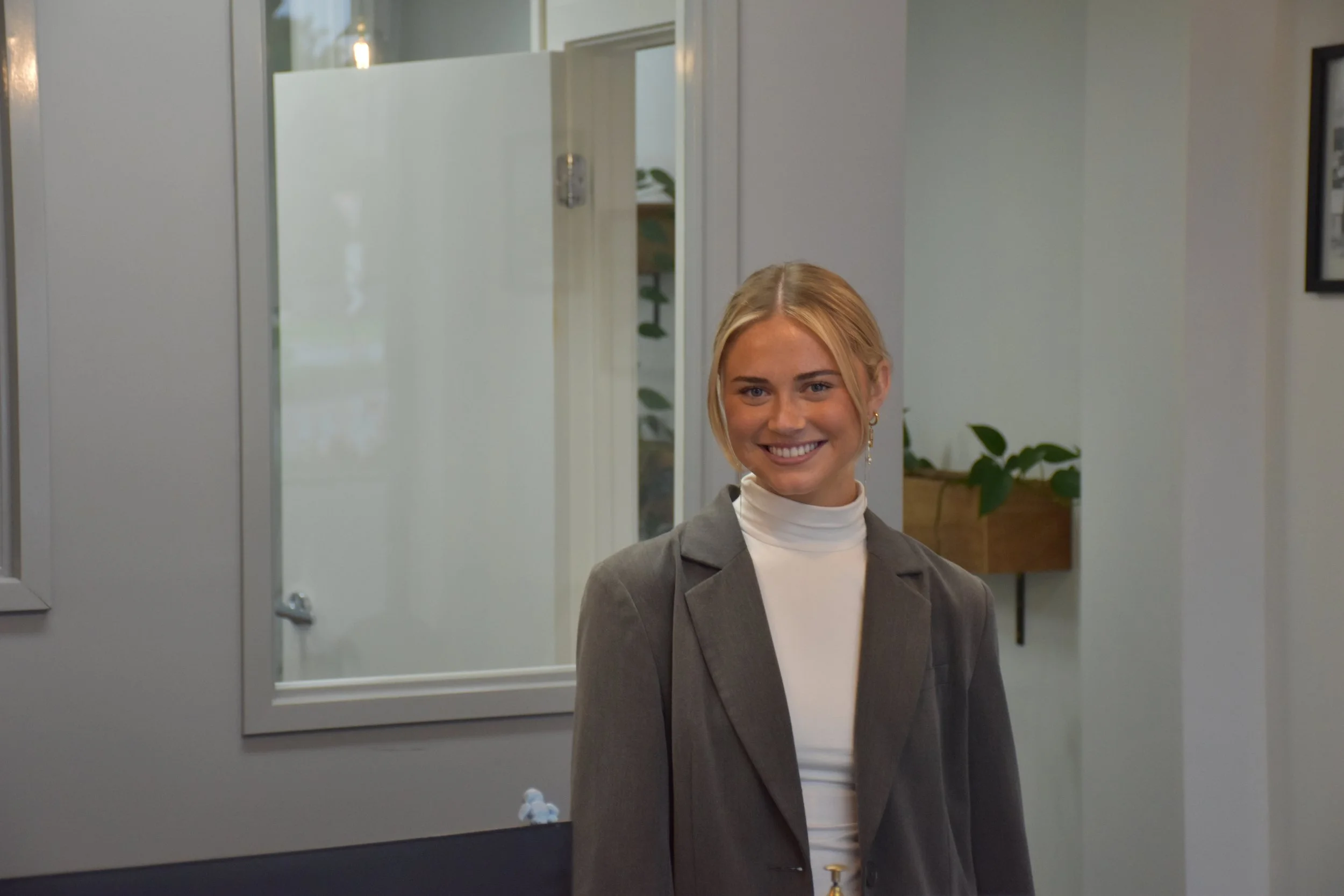 A young woman with blonde hair smiling in front of a mirror in a modern, minimalistic room. She is dressed in a gray blazer and white turtleneck and has earrings, with a plant visible in the background.