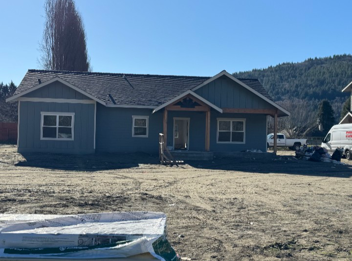 A newly constructed house with blue siding, a front porch, and driveway, surrounded by construction materials and vehicles, with a mountainous landscape in the background.