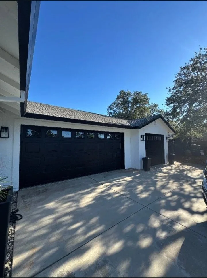 The image shows a driveway and garage of a house with two black garage doors, a black trash bin, and external lighting. There are shadows of trees cast on the concrete driveway, and the sky above is clear and blue with a few trees visible in the back