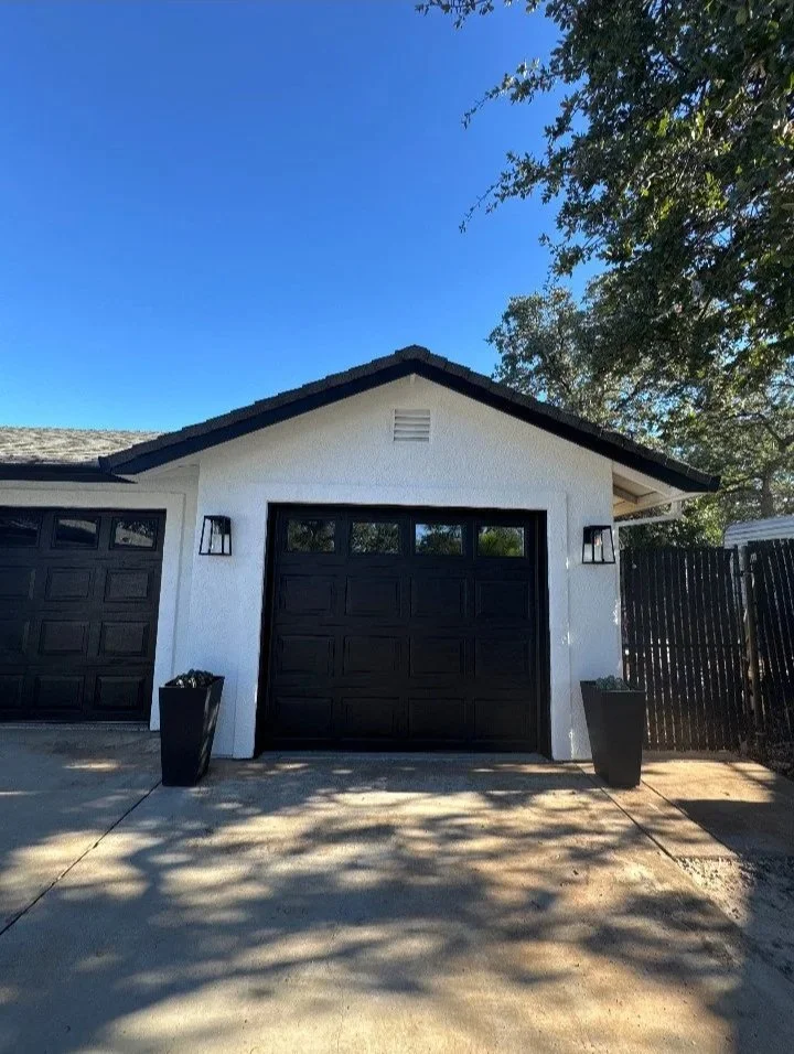 A white house garage with two black garage doors, black wall-mounted lights, and two large black planters, with a concrete driveway and shadows of trees, under a clear blue sky.