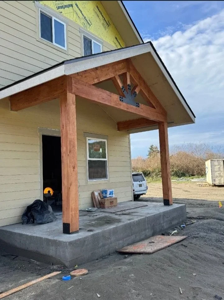 A house under construction with a new front porch and wooden support beams. The house is beige with white trim, and there are construction materials and tools on the porch and ground.