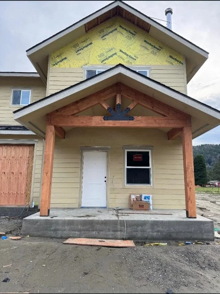 The front of a house under construction with a new porch and exterior walls in progress, showing yellow insulation and unfinished wood framework.