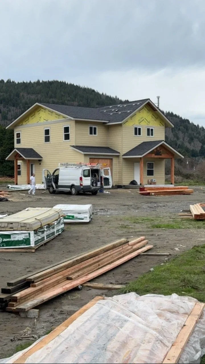 Under construction two-story house with yellow siding, black roof, and wooden porch, surrounded by building materials and a van, with mountains in the background.