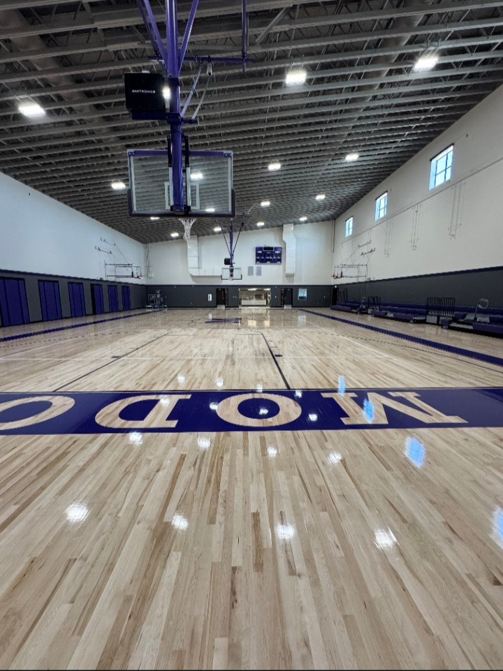 Empty basketball court with wooden flooring, purple and black colors, basketball hoops, and benches along the walls.