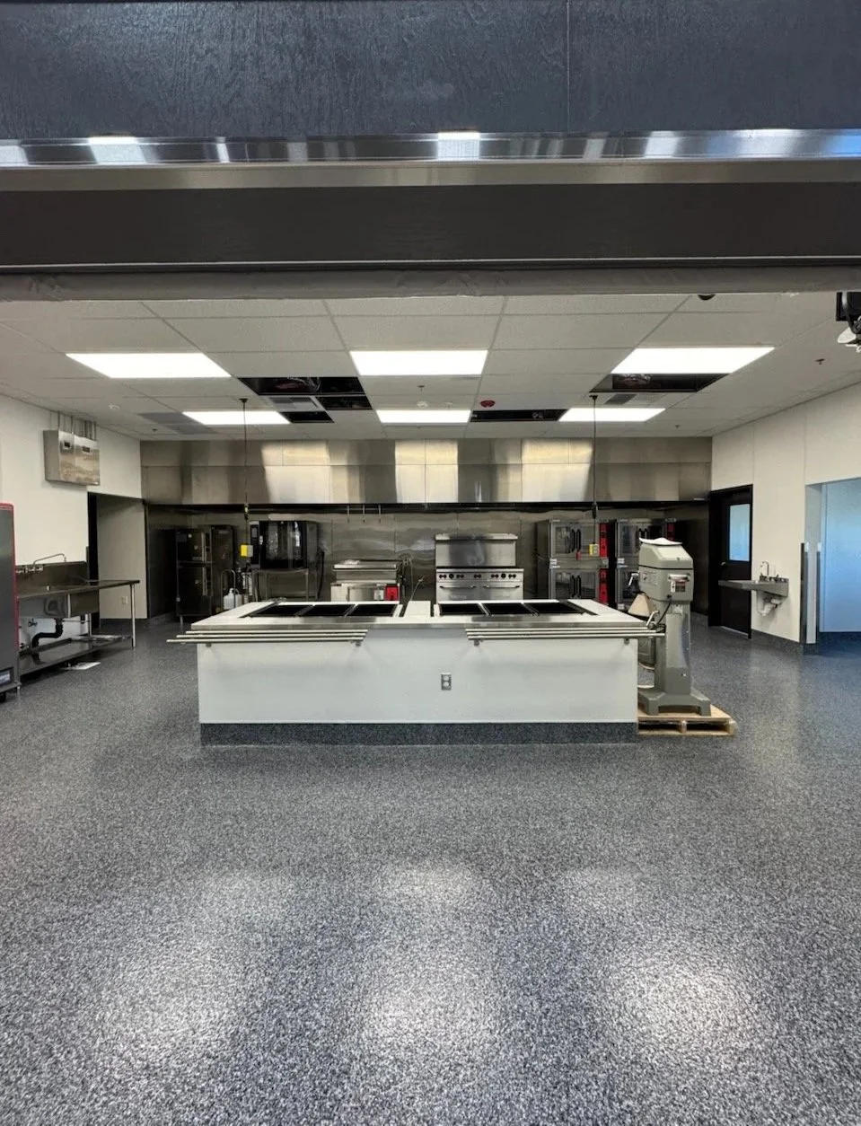 Empty commercial kitchen with stainless steel appliances and black speckled floor.