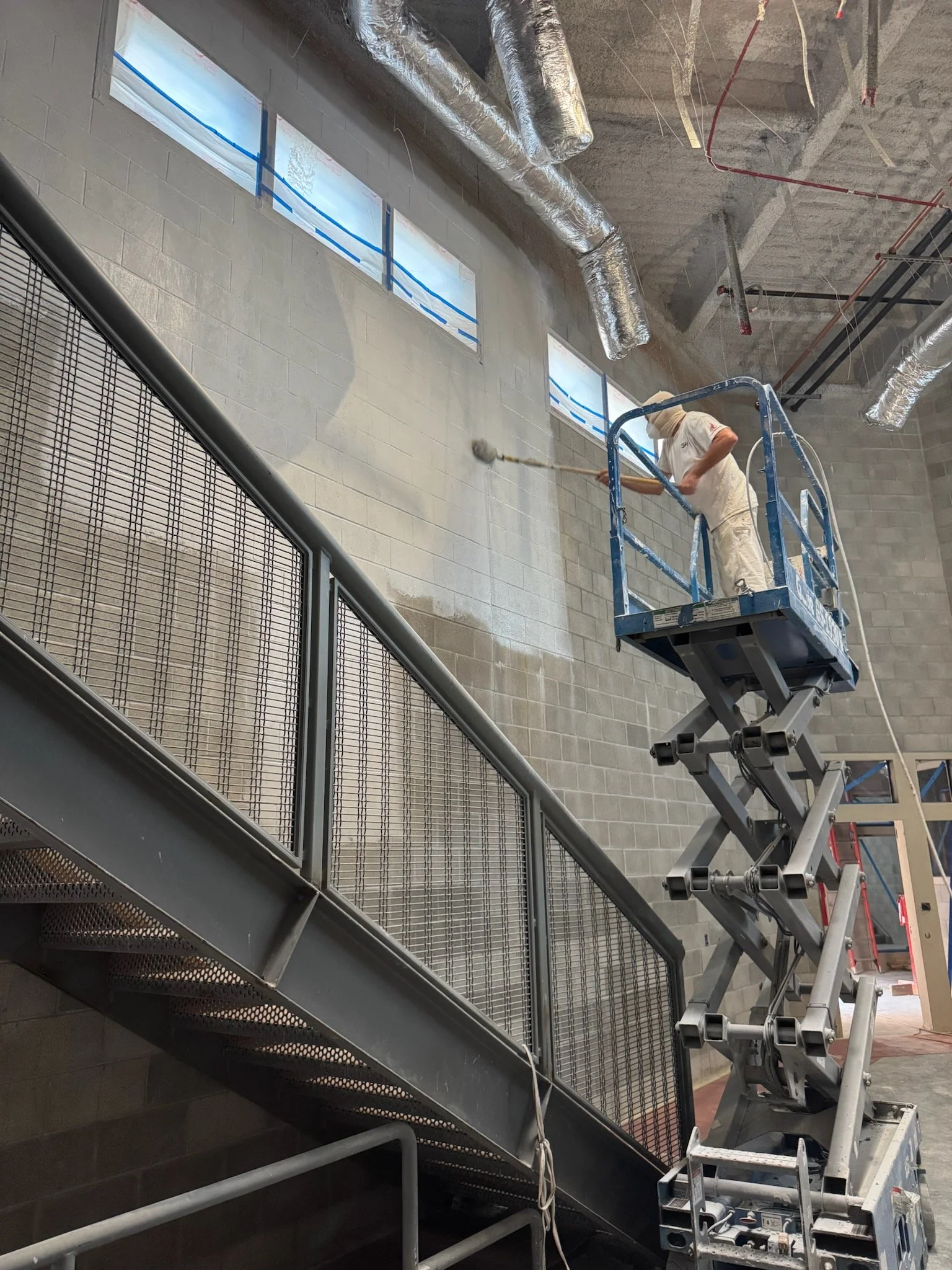 A worker operating a scissor lift is painting a high wall with a roller brush. The wall has three small windows near the ceiling, and there are large silver ducts and exposed pipes on the ceiling. The site appears to be a construction or renovation project.