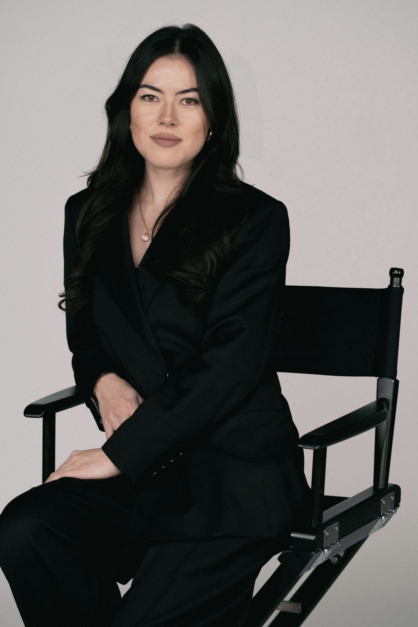 A woman with long black hair wearing a black blazer and gold jewelry sitting on a director's chair against a plain light gray background.
