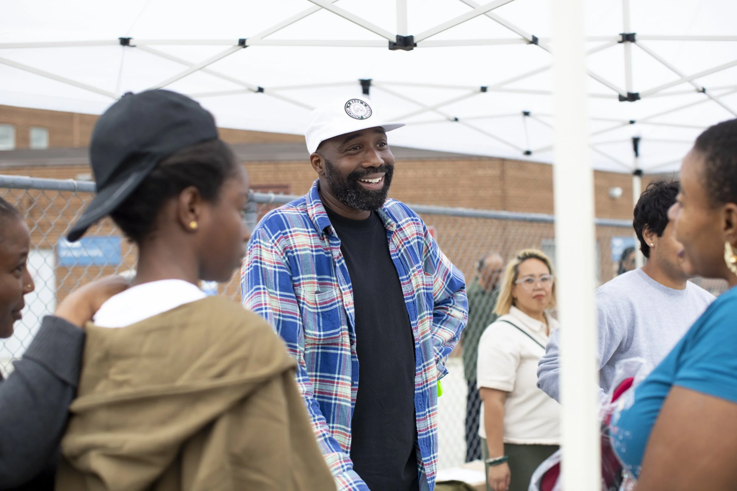 A group of people gathered outdoors under a white canopy, with a man in a white cap and plaid shirt smiling in the center of the group.