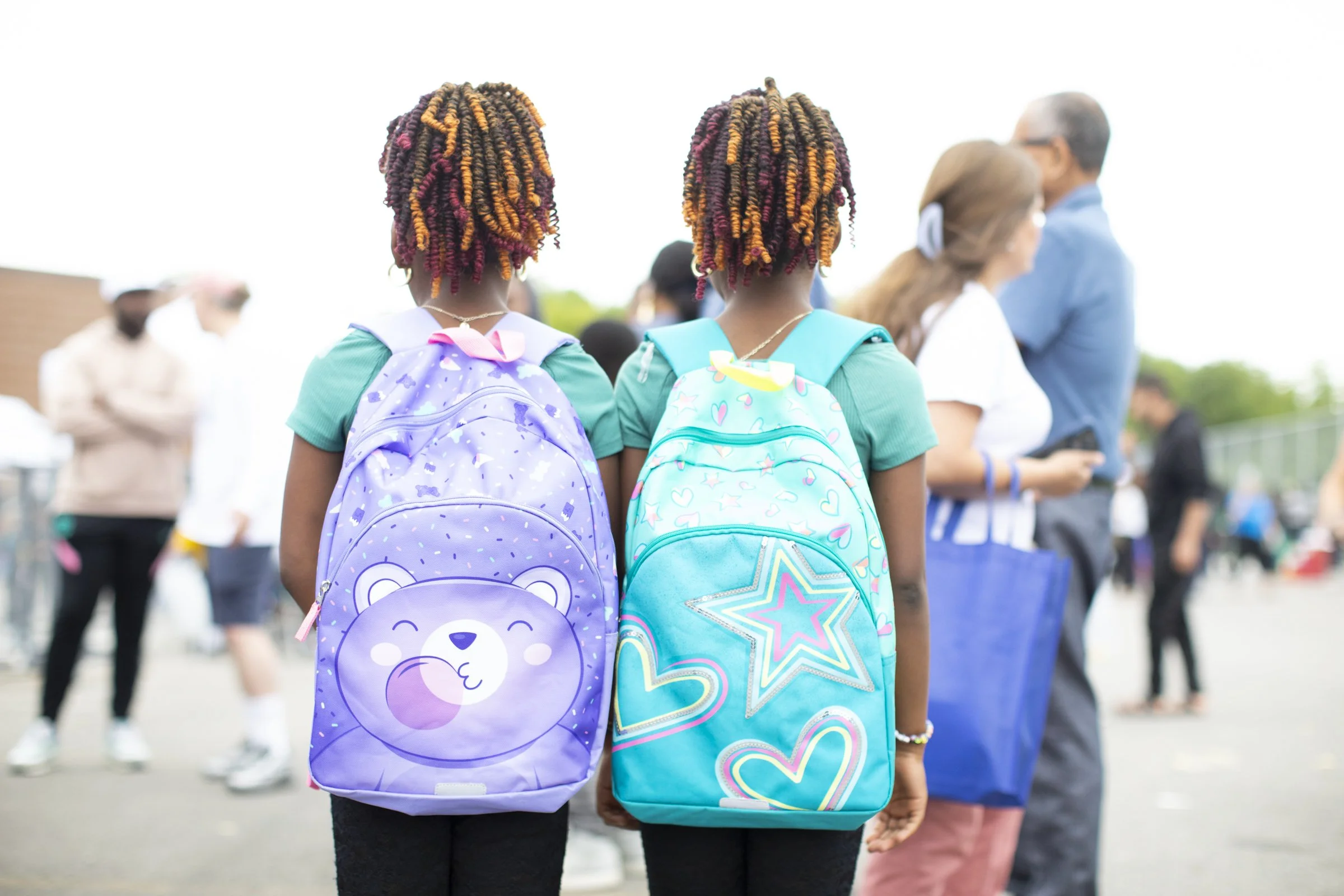 Two girls with colorful backpacks standing outdoors, with blurred adults in the background.