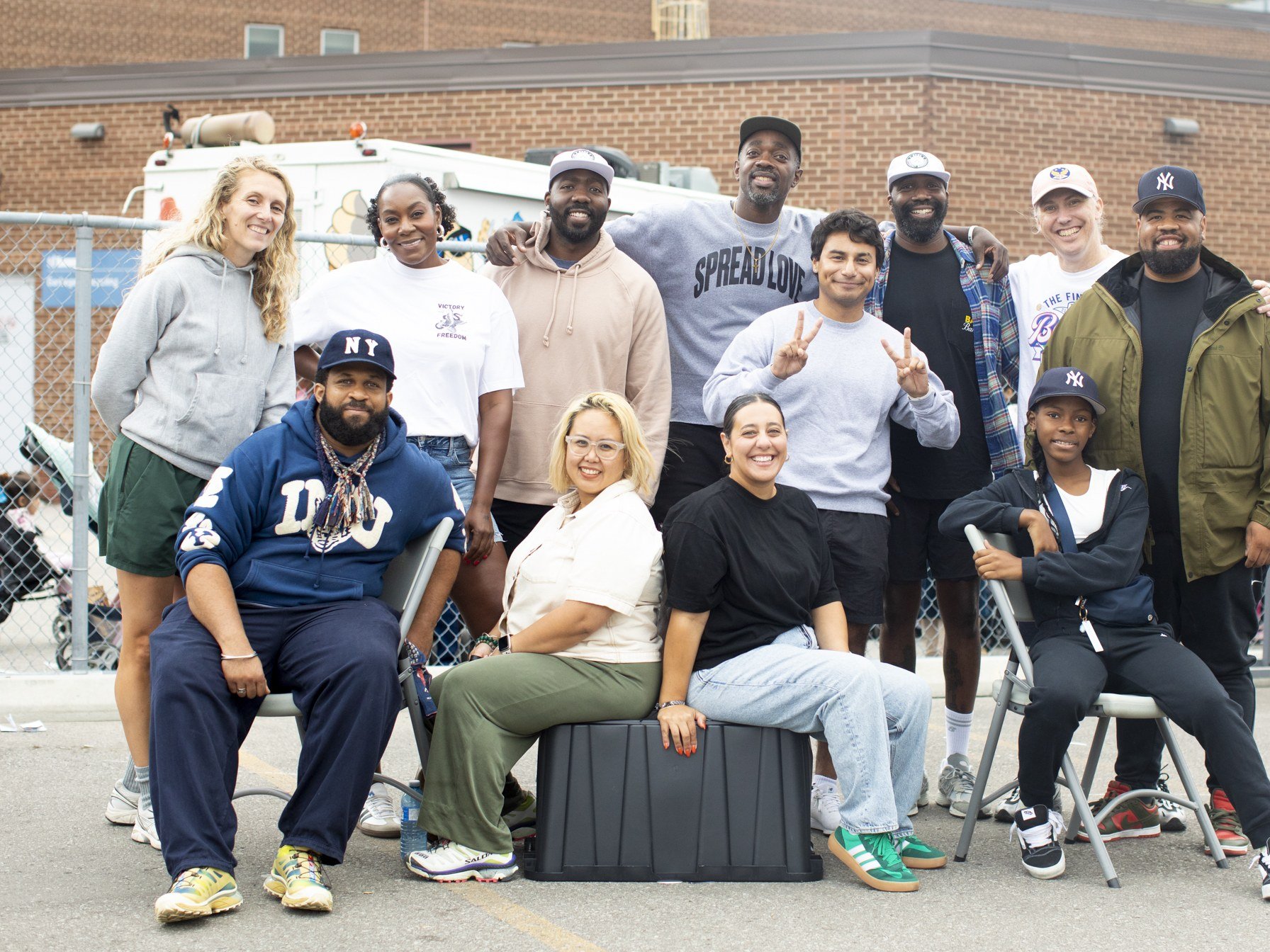 Group of 12 diverse people posing outdoors in front of a brick building and chain-link fence, some sitting and some standing, with arms around each other, smiling for the camera.