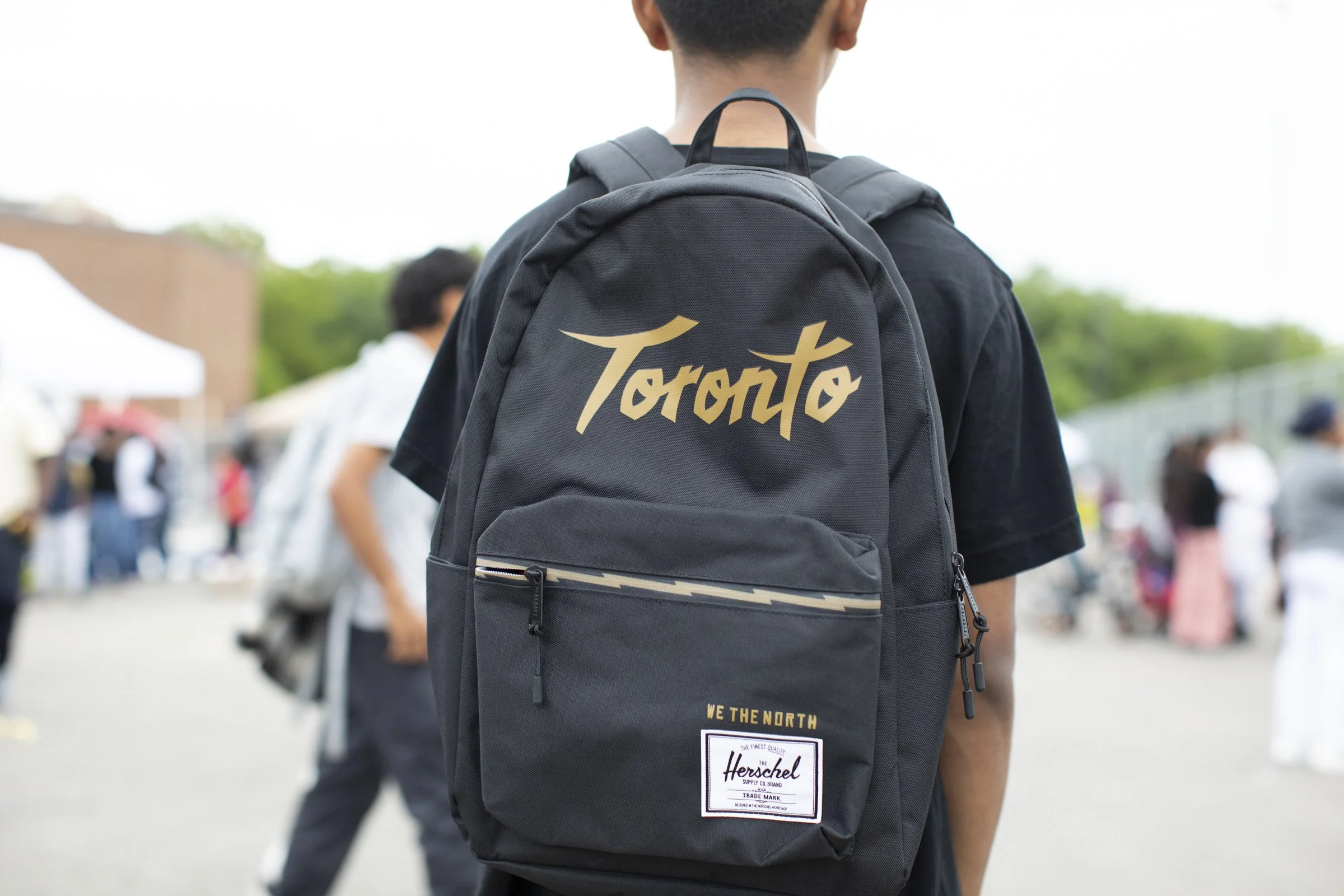 Person wearing a black backpack with the word 'Toronto' written on it, standing outdoors with other people in the background.