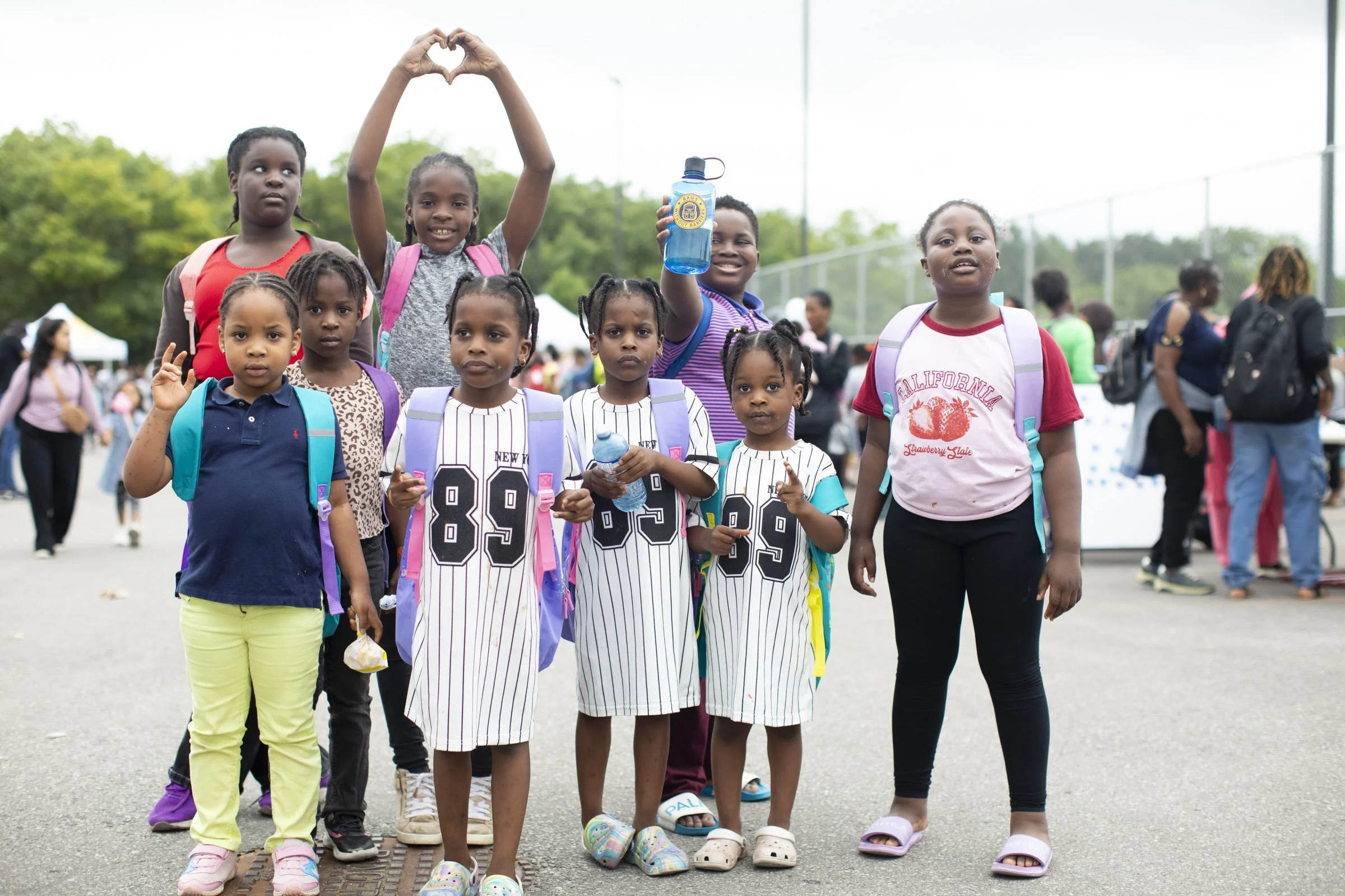 A group of young children, mostly girls, standing outdoors on a cloudy day. Some are holding water bottles, and a few are making heart shapes with their hands. They are wearing casual clothing and backpacks, and there are other people and trees in the background.