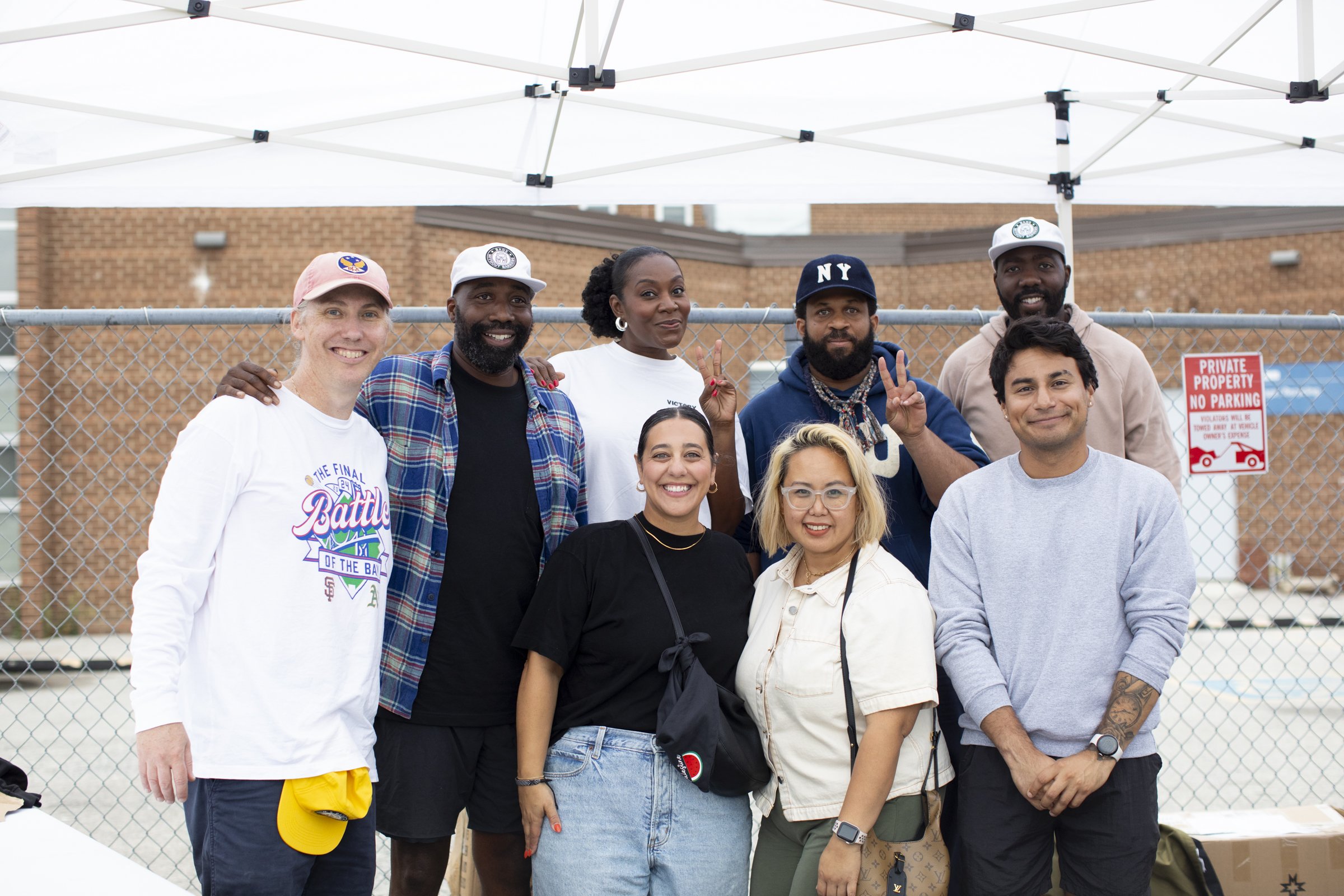 Group of nine diverse people smiling in front of a chain-link fence outdoors, with tents overhead and a sign that reads 'Private Property No Parking' in the background.