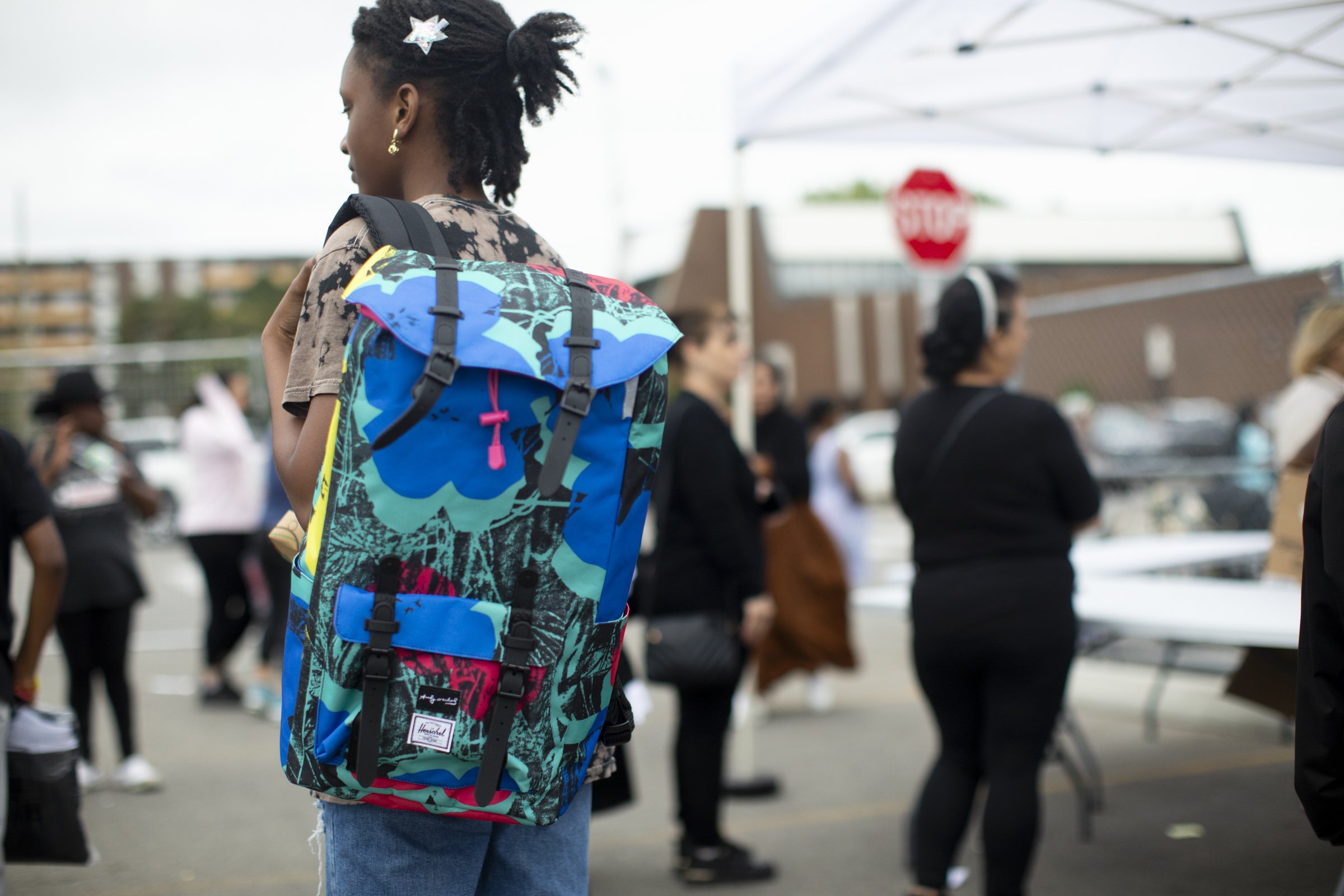 A young woman with a backpack standing among a crowd of people outdoors, near a stop sign and a white canopy.