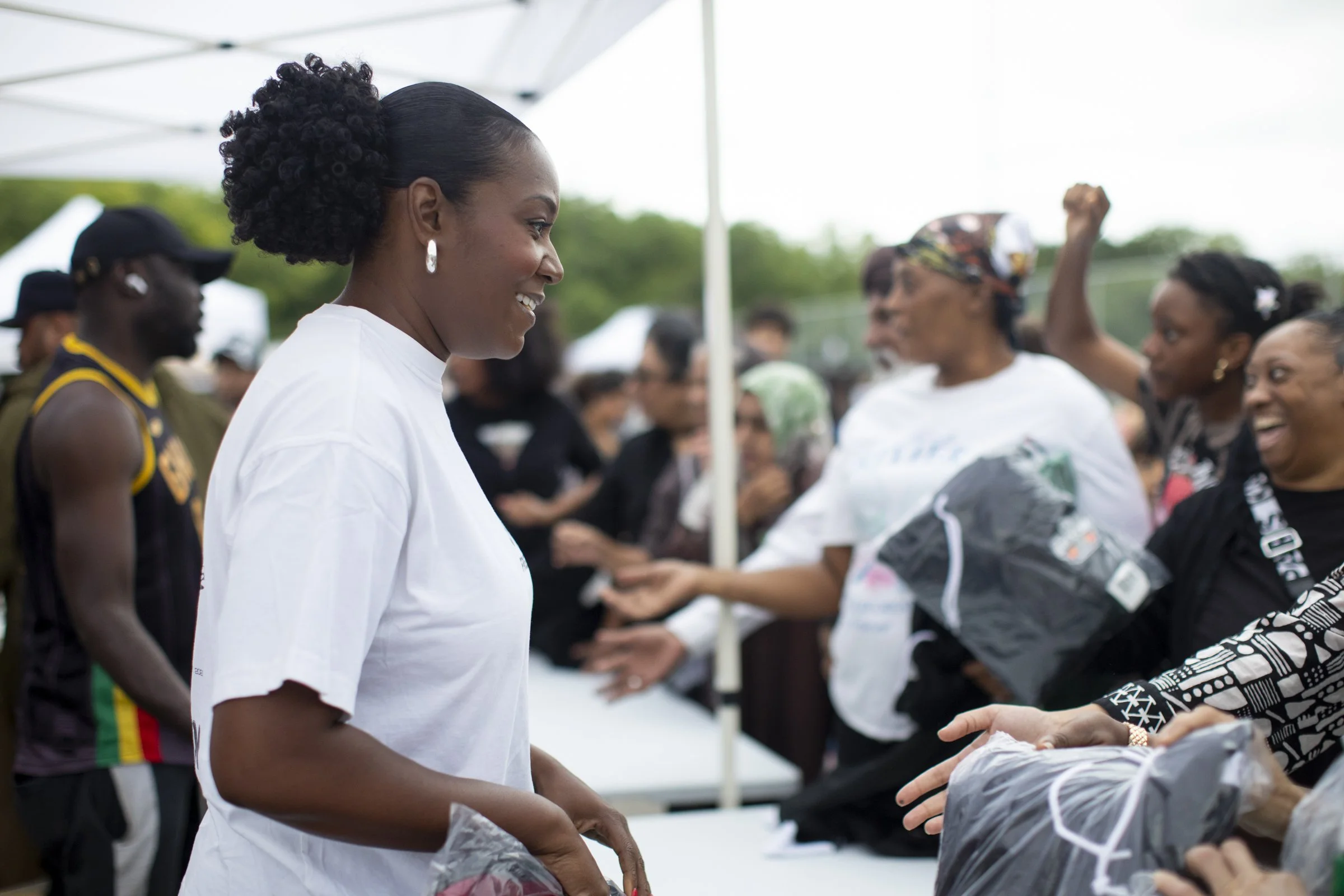 A woman with curly hair in a bun wearing earrings is smiling and shaking hands with another person at an outdoor event. There are several people in the background, some wearing black shirts and others in casual attire, under a white tent canopy.