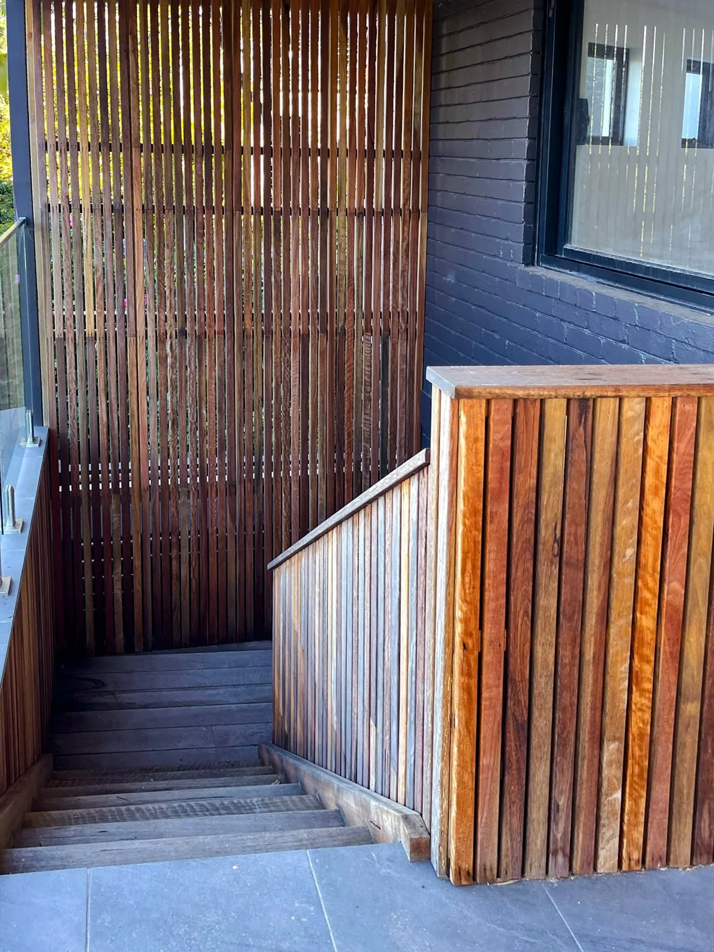An outdoor staircase with wooden handrails and vertical wooden slats, next to a black brick wall with a window.