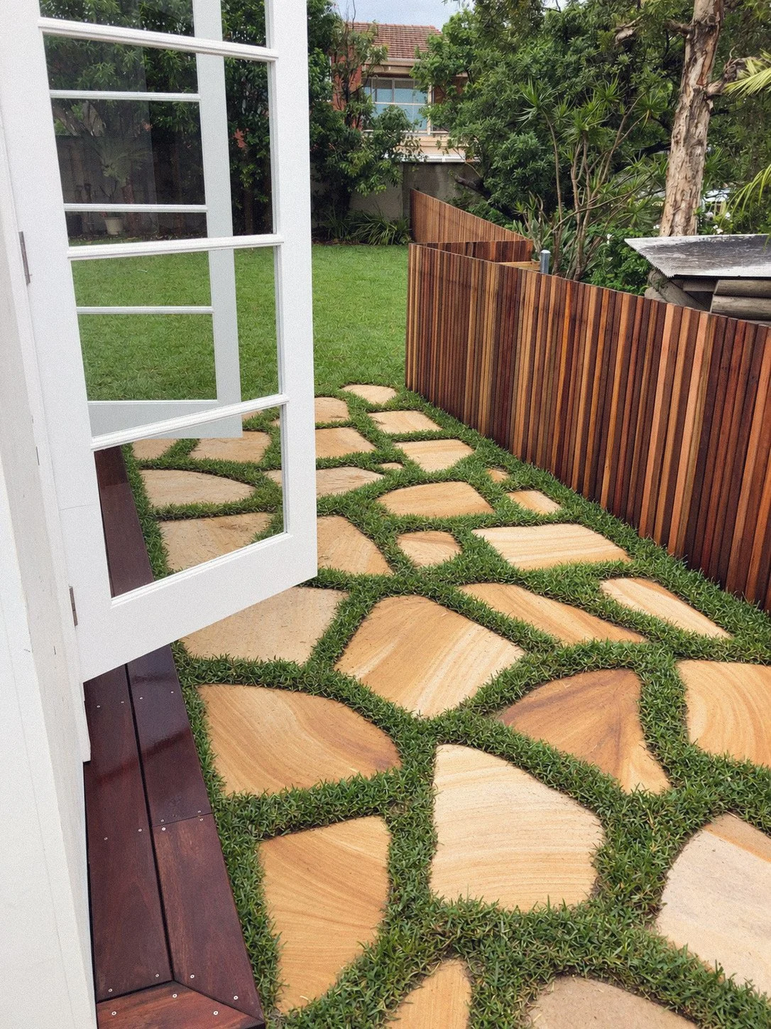 View of a backyard patio with geometric stone pathway bordered by grass, a wooden privacy fence, and lush greenery including trees and shrubs. An open French door leads outside.