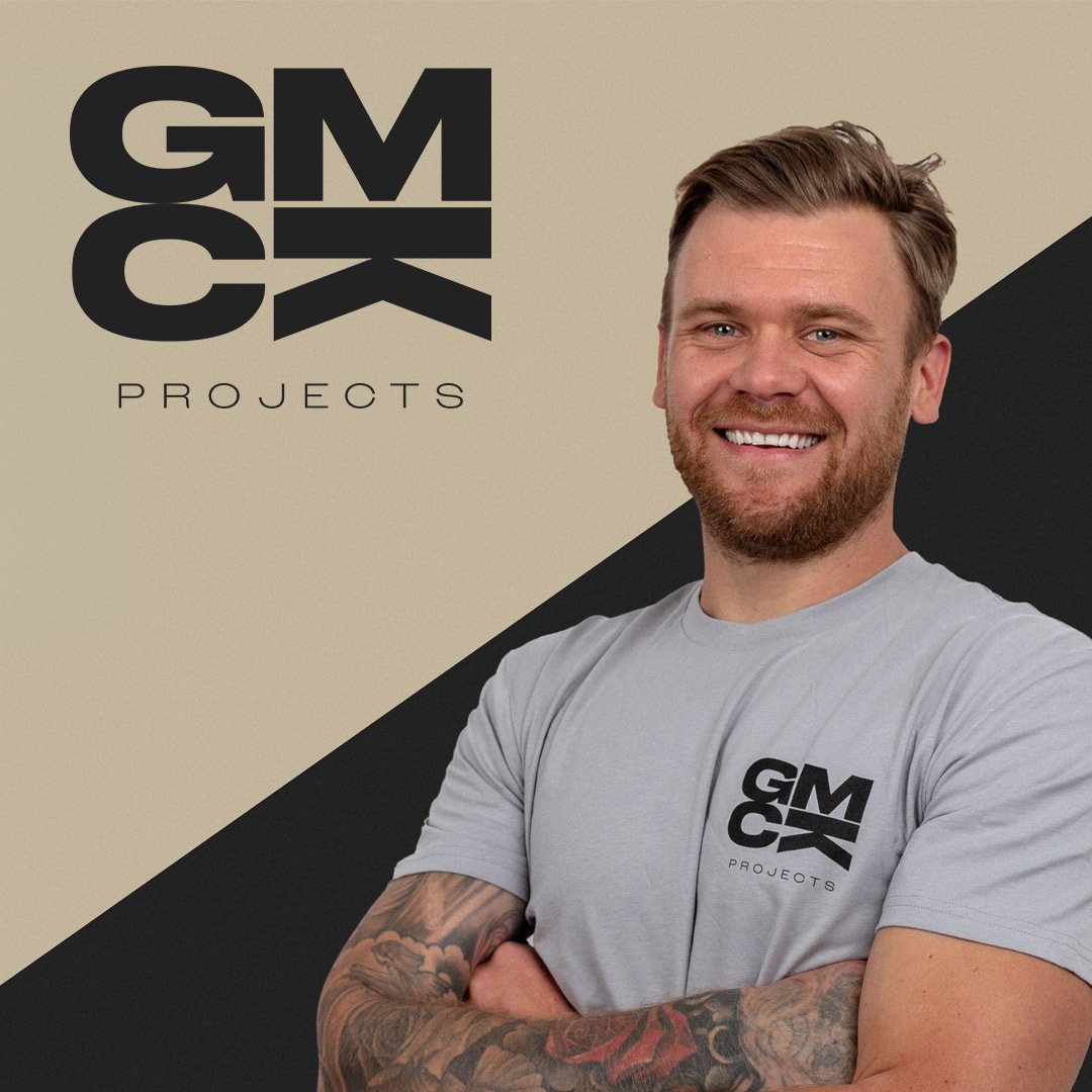 A smiling man with tattoos on his arms wearing a light gray t-shirt with a black and white logo 'GMCX Projects' on it, standing in front of a beige and black diagonal split background with the same logo and company name.