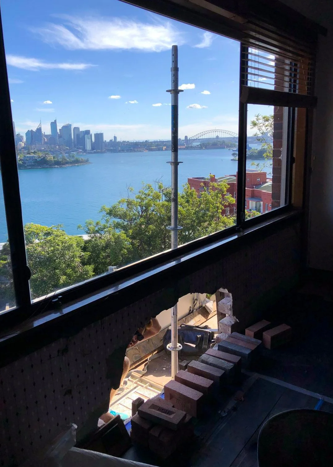 Looking out a window at the Sydney Harbour skyline with buildings, a bridge, trees, and water, with construction work inside.