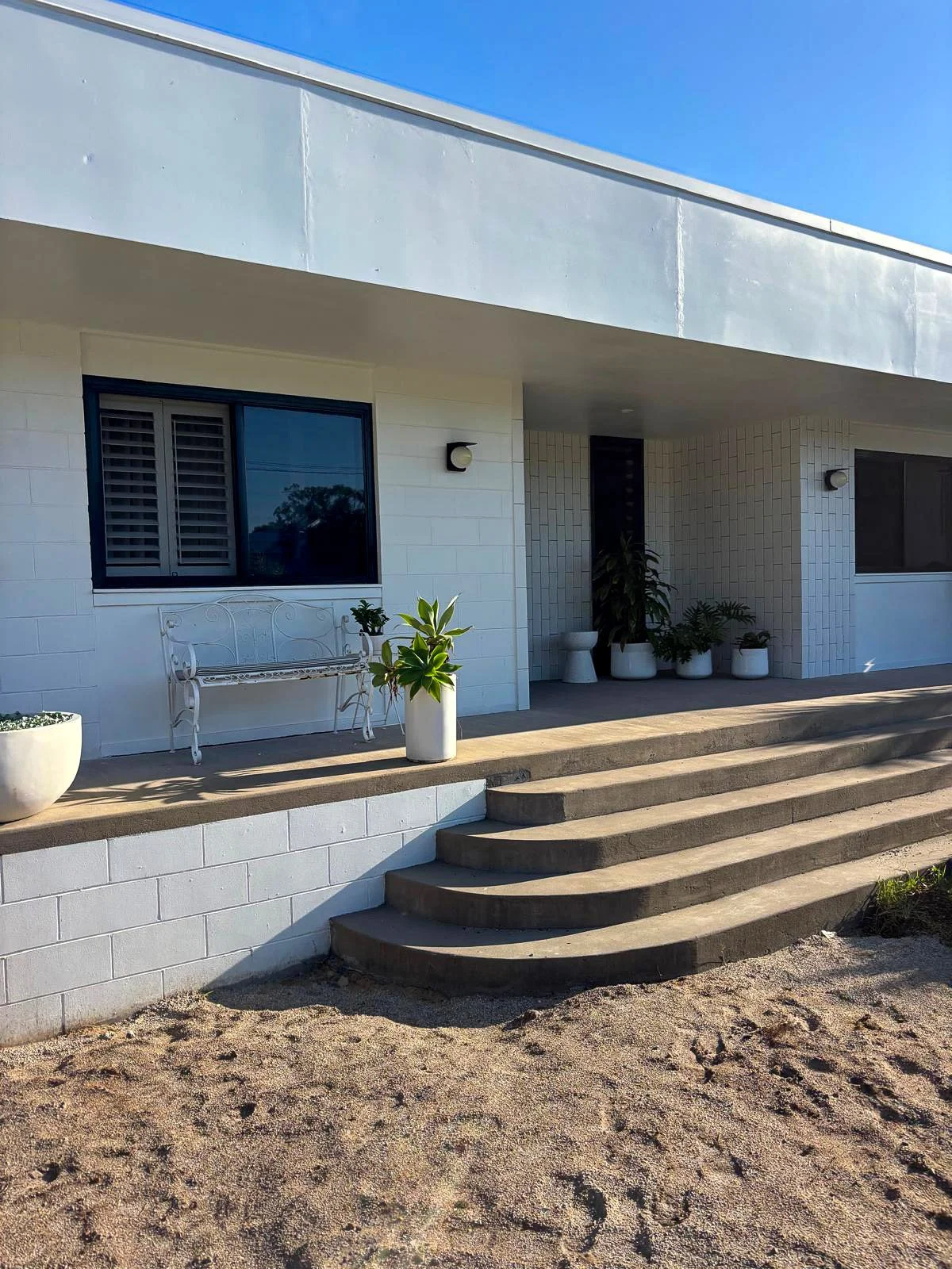 Front porch of a modern house with white exterior, concrete steps, and potted plants, under a bright blue sky.