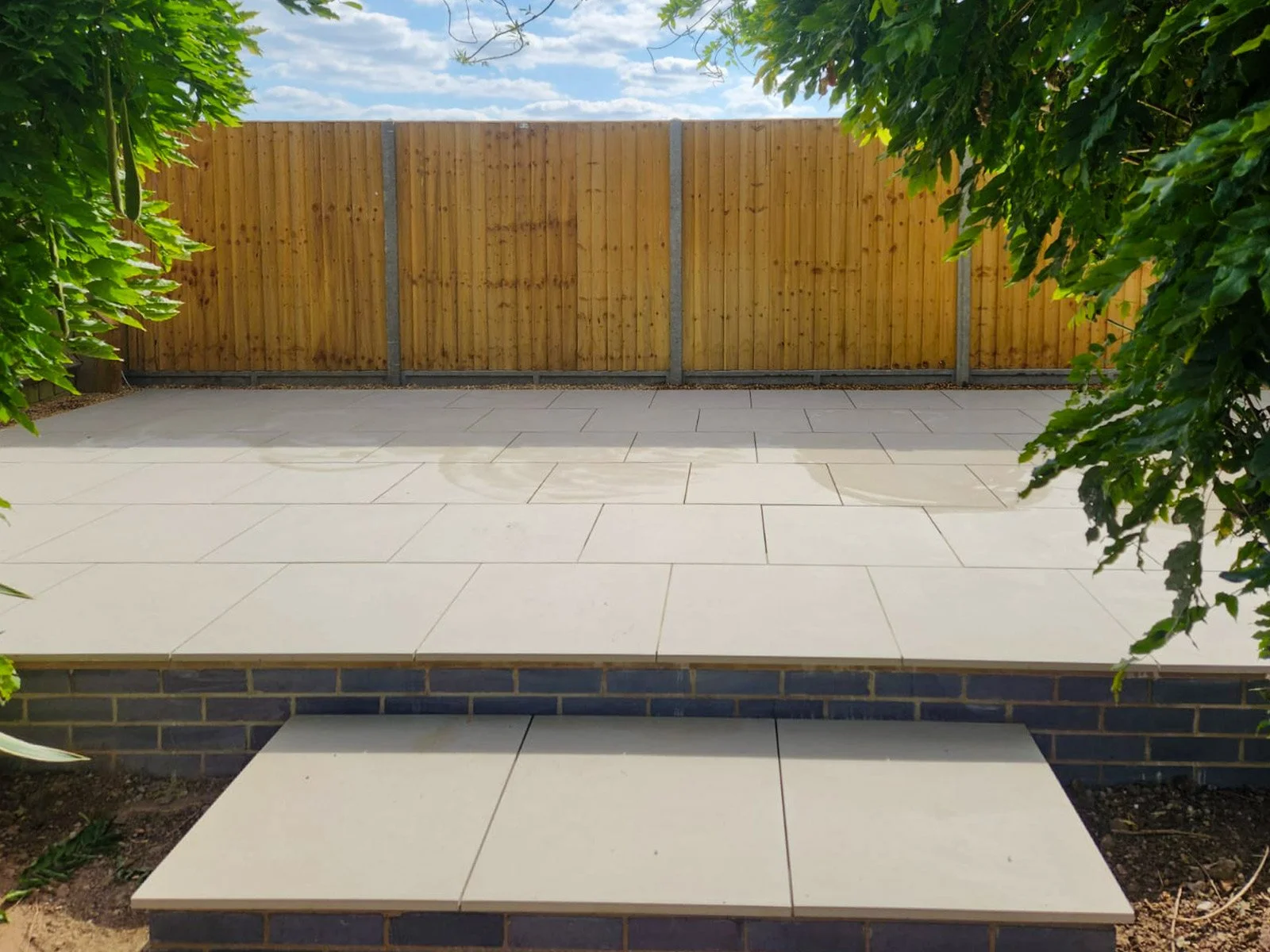 A backyard patio with beige tiles, a wooden privacy fence, and greenery on both sides.