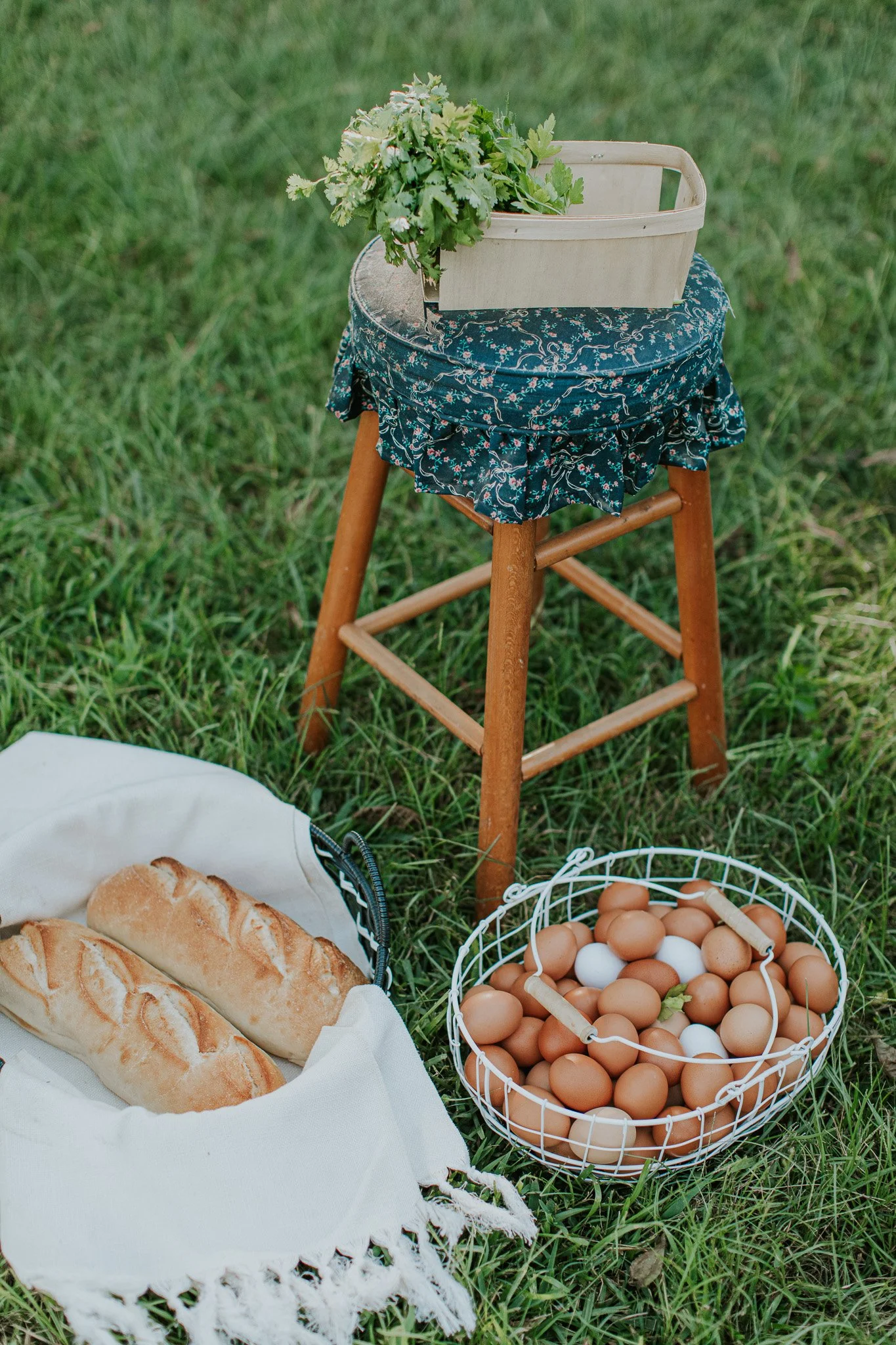 A picnic scene with a basket of eggs, baguettes on a white cloth, and a wooden basket with greens on a small stool covered with a dark blue floral cloth, set on grass.
