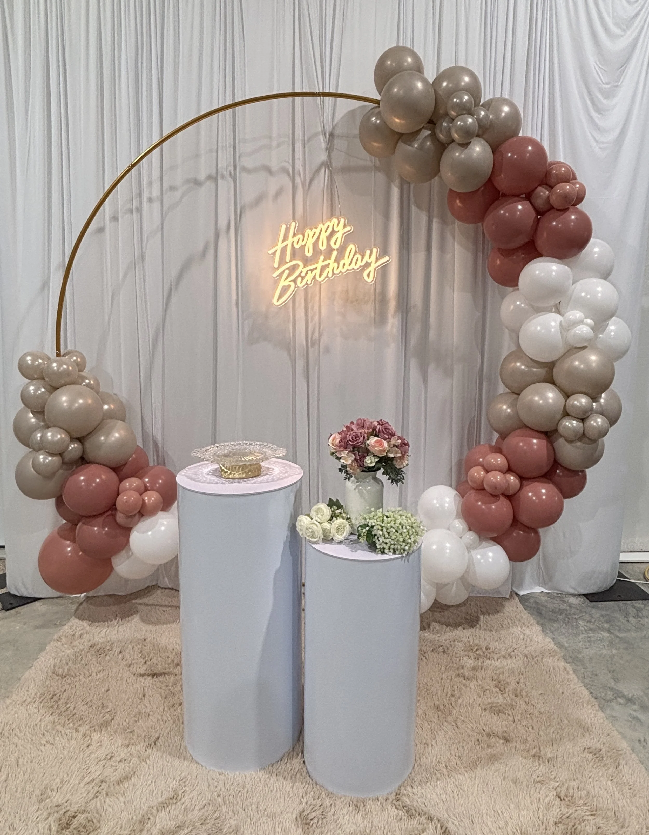 Decorative birthday display with a balloon arch in shades of beige, pink, and white, a neon "Happy Birthday" sign, and tables with flowers and cake on a beige carpeted floor with a white curtain backdrop. Perfect for you next event in Gulfport, MS.