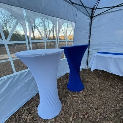 Two tall cocktail tables, one with a white cover and the other with a blue cover, in a white event tent outdoors with trees visible through the windows.