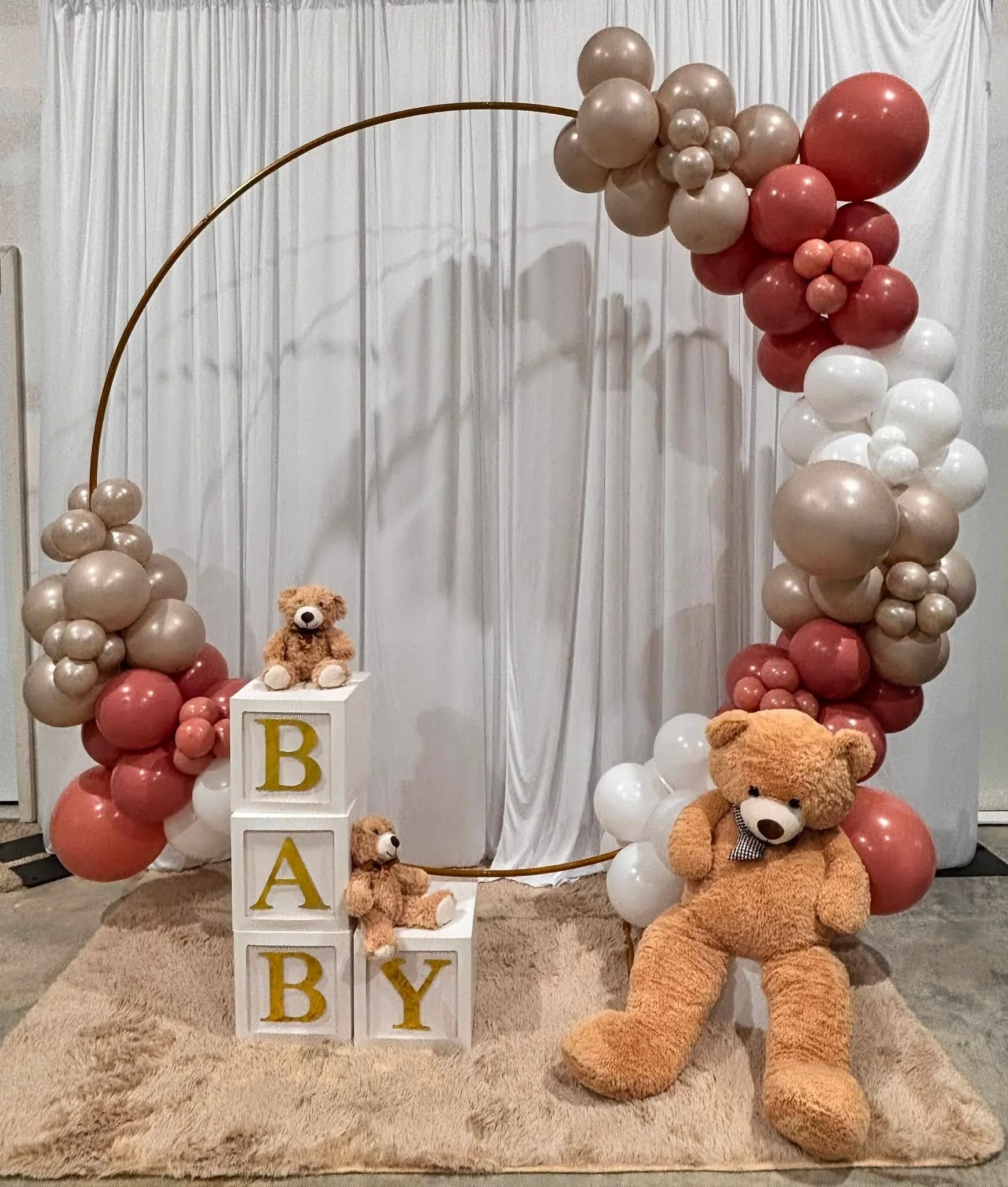 A teddy bear setup with balloon arch, blocks with letters spelling baby, and a white curtain background for a baby shower or celebration.