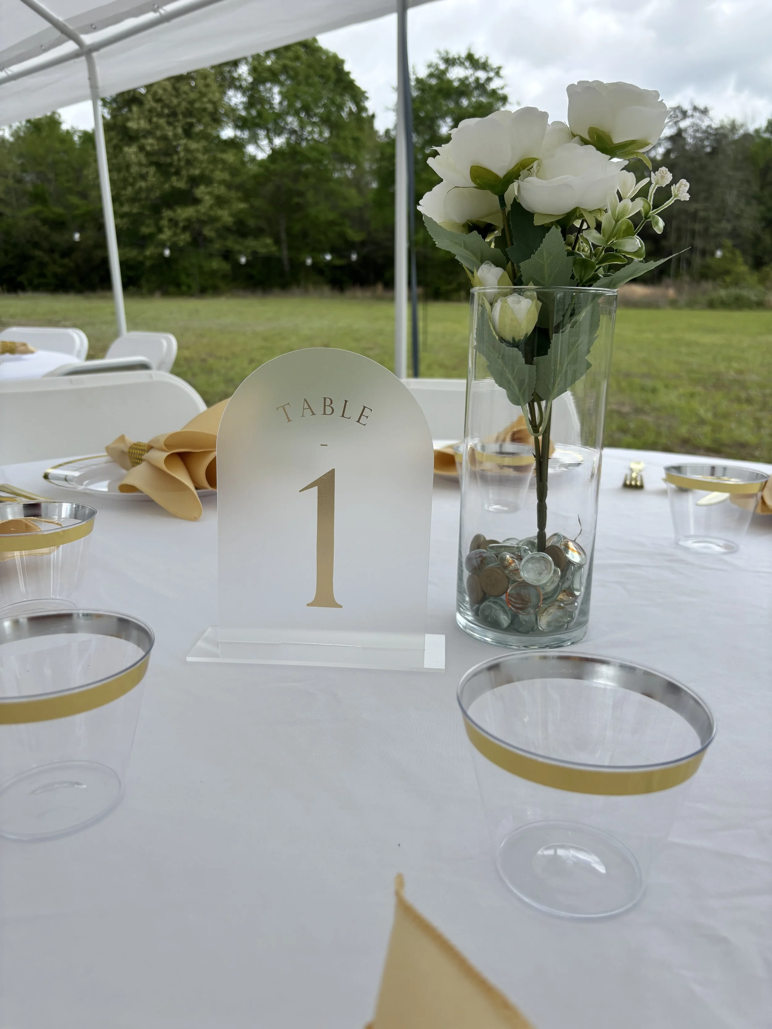 A white table set for an event outdoors with a white canopy overhead. The table has a centerpiece with white flowers in a clear glass vase filled with decorative stones, a table number card with the number '1', and empty clear cups with gold rims.