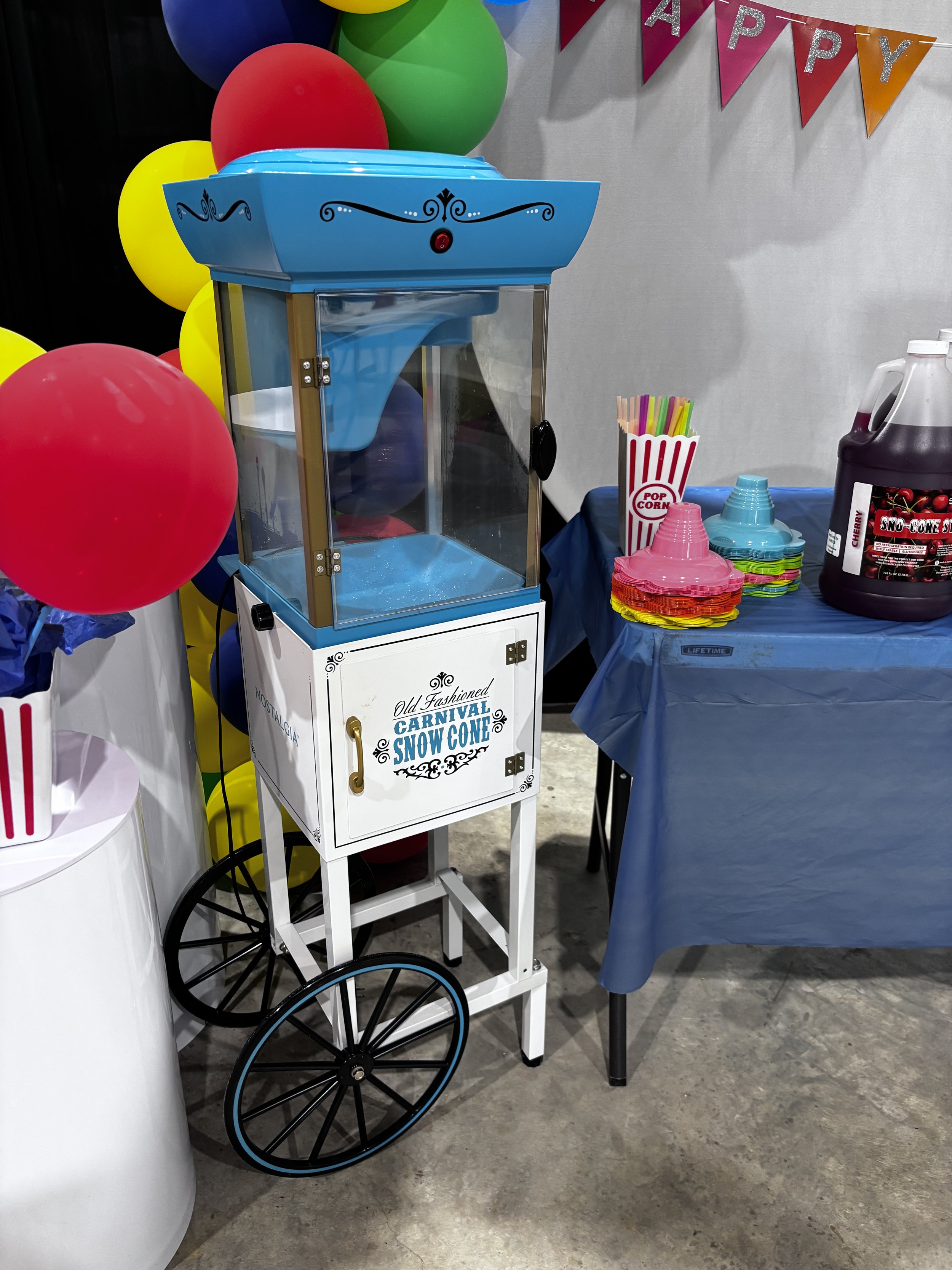 A snow cone machine with a white base and blue top, decorated with black swirl designs, and a clear window showing snow inside. Next to it are colorful paper cups and straws on a table covered with a dark blue cloth. The background features balloons in red, yellow, green, and blue, and a banner with pink and orange triangular flags spelling 'HAPPY' in glittery silver letters, hanging on a wall.