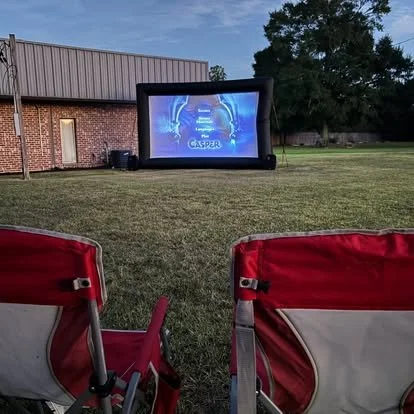 Outdoor movie screening setup with an inflatable screen showing a film, two red chairs facing the screen, on a grassy area near a brick building.