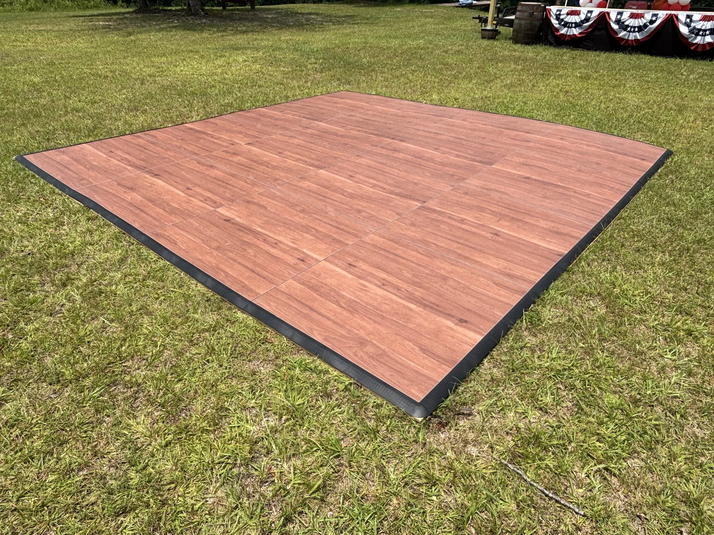 A wooden dance floor on grass at an outdoor event with patriotic red, white, and blue bunting on a stage in the background.