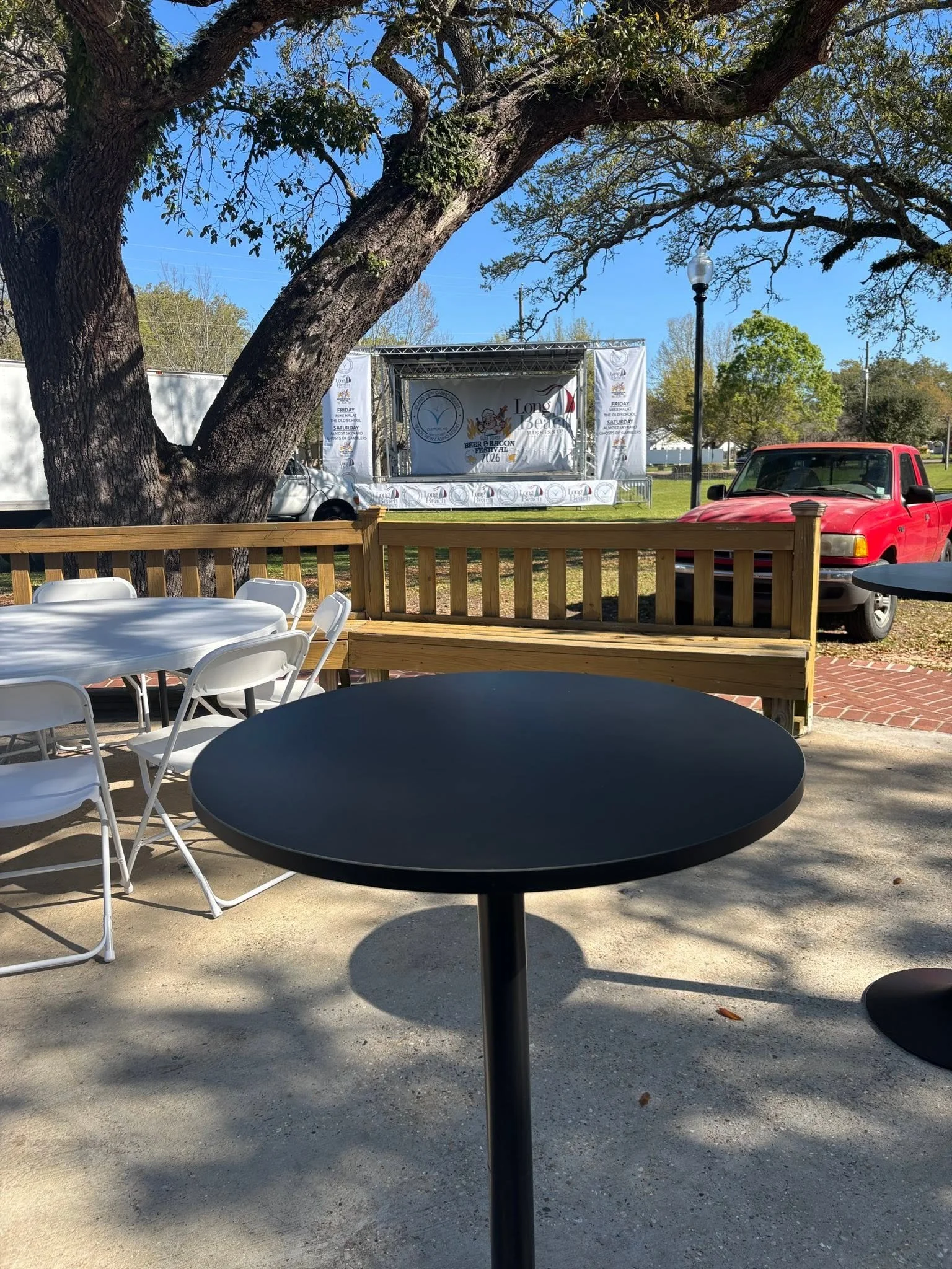 Empty outdoor seating area with white and black tables, a wooden fence, and a stage setup in the background for an event, underneath large trees with a blue sky overhead.